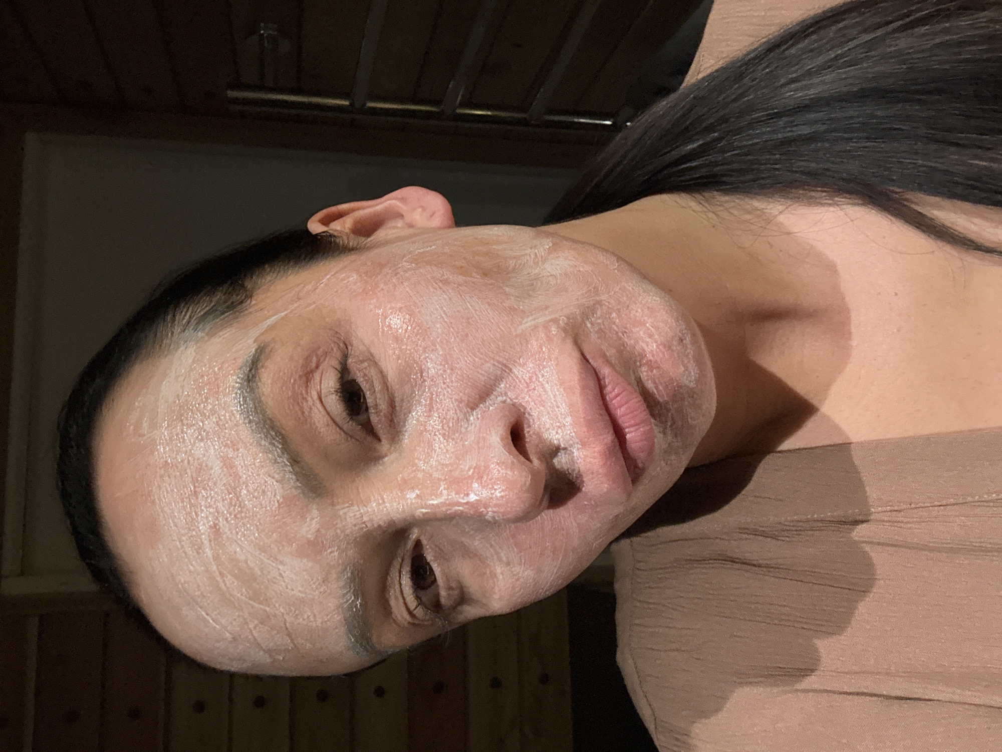 A close-up, angled shot of a woman with dark hair and a white facial mask applied to her face. The woman is wearing a light brown or nude-colored top. The background shows wooden walls or paneling. The location and city of Chaikovsky, Russia cannot be determined from the visual information in the image.