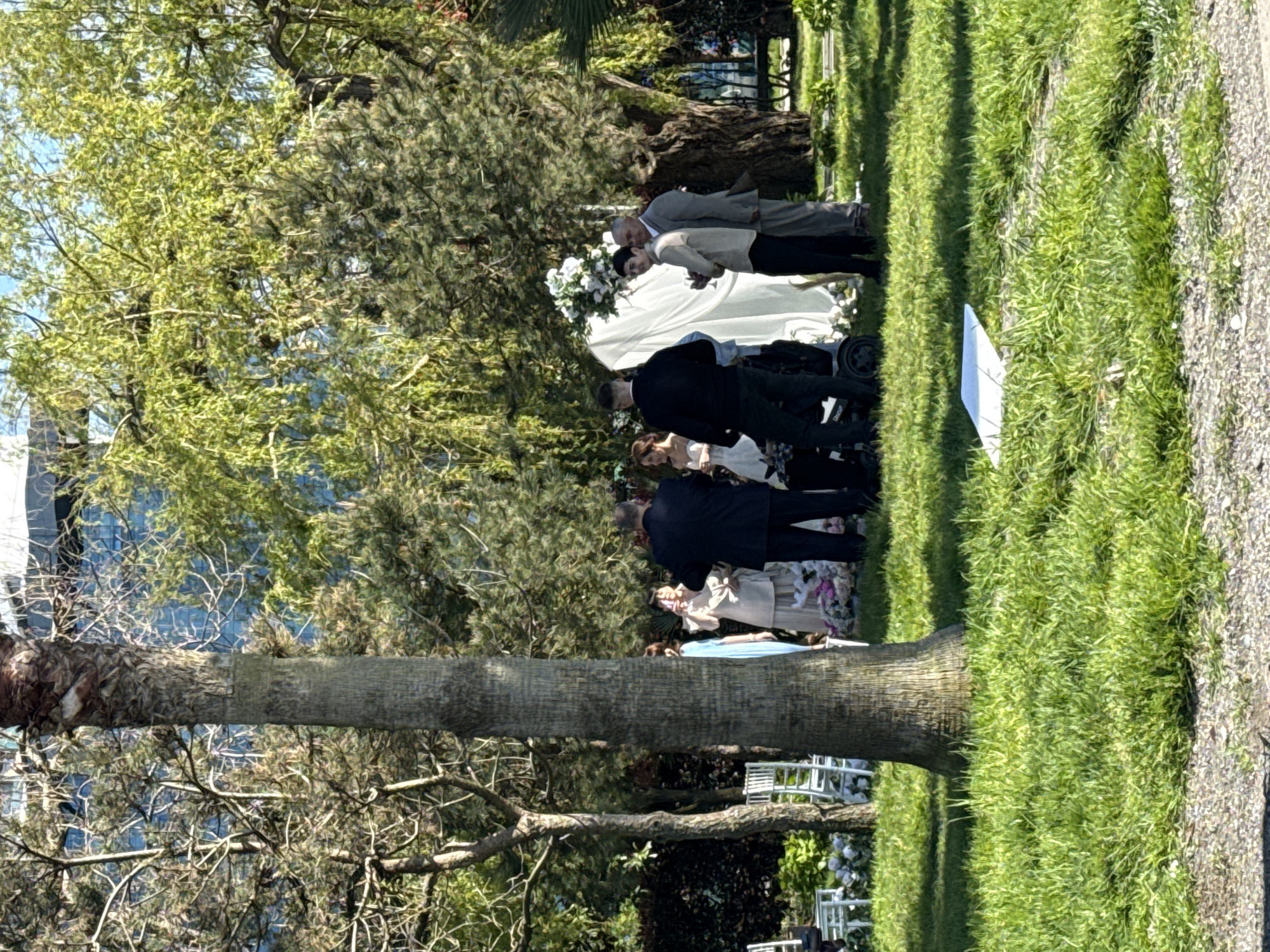 A wedding ceremony is taking place outdoors in Batumi, Georgia. The location appears to be a garden or park setting. There are at least a dozen people attending. There are trees, foliage, chairs, and a wedding altar adorned with white flowers. The ceremony is taking place on a grass lawn next to a stone path.