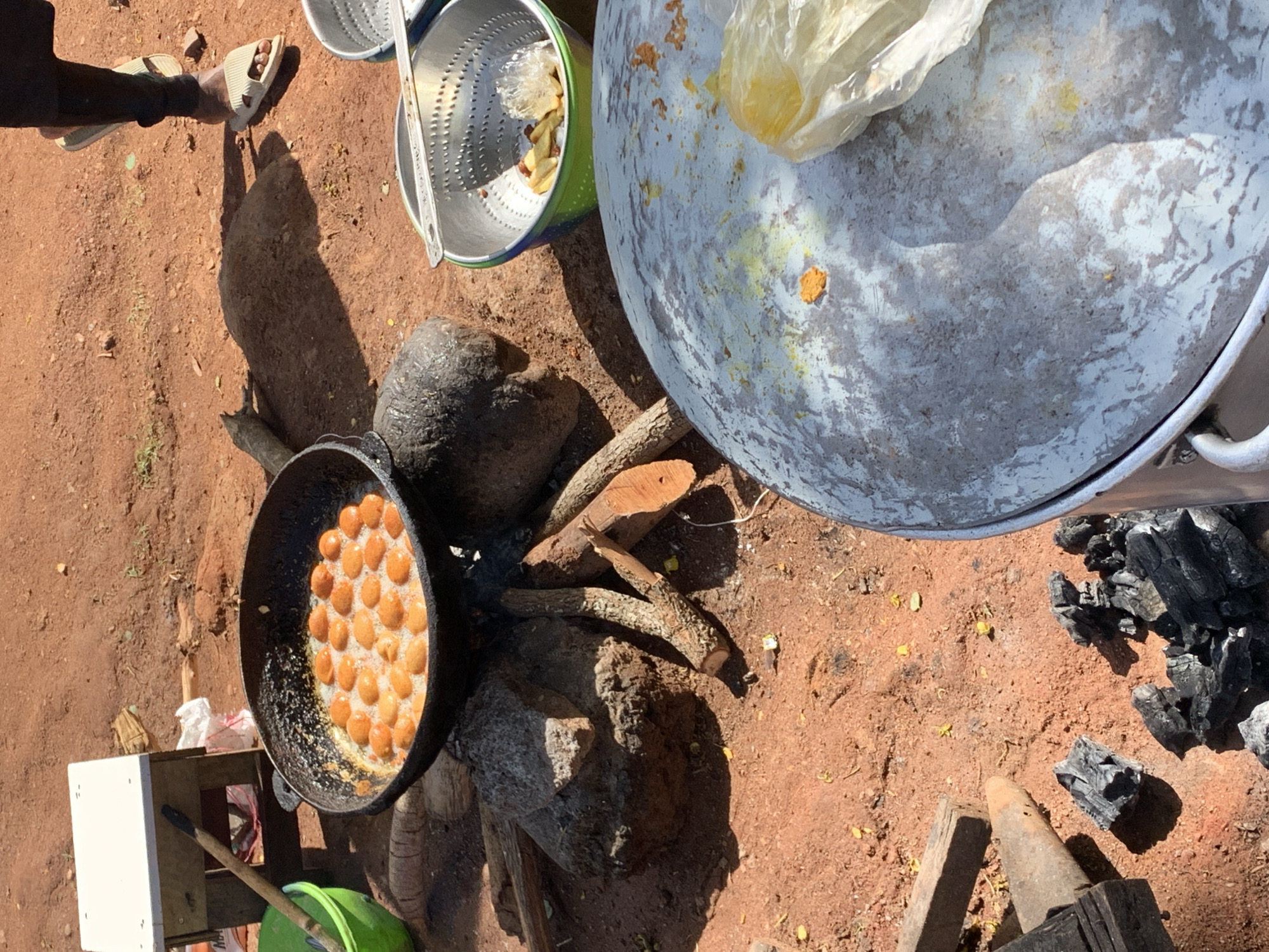 An overhead photograph captures an outdoor cooking scene on reddish-brown earth, likely in Kabong, Nigeria. The strong, defined shadows suggest it is midday or early afternoon under bright, sunny weather conditions.
In the upper left, the lower leg and foot of a person wearing a light-colored sandal are visible, casting a long shadow onto the dusty ground.
The central activity revolves around a large, black, round pot, possibly cast iron, filled with numerous small, round, golden-brown dough balls frying in bubbling oil. This pot is positioned over an open fire, supported by several rocks and pieces of firewood, indicating a traditional cooking method. Remnants of charred wood and ash are visible around the cooking area.
To the right of the cooking pot, a large, light-colored, round lid or container, which appears to be metallic or painted, rests on the ground. It has a handle and shows yellow stains and a clear plastic bag on its surface. Further to the right, a heap of black charcoal pieces is scattered on the dry earth.
In the upper central part of the image, a shiny metal colander or strainer, possibly placed within a green bucket, contains some light-colored food scraps and another clear plastic bag. A small white box and a wooden stick are also present near the cooking setup. The scene depicts a functional, rustic outdoor kitchen for preparing food, possibly street snacks like puff-puffs, in an arid environment. No discernible text is visible.