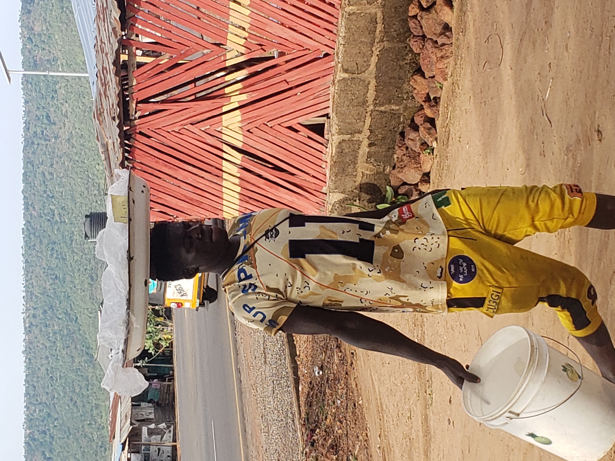 A man walks on a dirt shoulder adjacent to a paved road in Adankolo, Nigeria. He is wearing a light yellow camouflage patterned shirt with the number "1" on the front, yellow shorts, and no shoes. On his head, he carries a rectangular white object covered in white plastic. In his right hand, he holds the lid of a white plastic bucket. In the background, a red-roofed building sits next to a street with a yellow vehicle and other structures. A lush green hillside rises beyond the buildings.