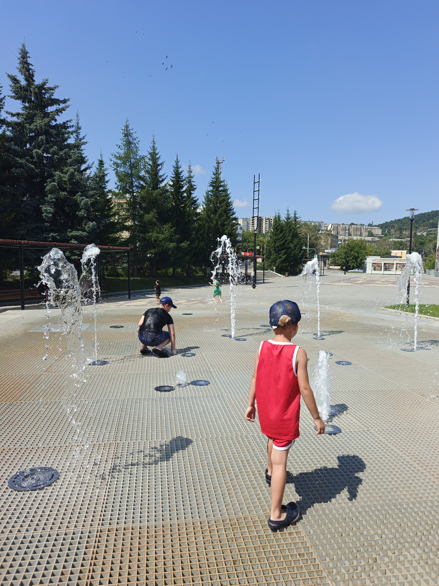 This image captures a sunny day scene at a public fountain plaza in Zlatoust, Russia. In the foreground, two young boys are interacting with the water jets. One boy, wearing a red tank top and red shorts, faces away from the camera, looking towards the fountains. He is wearing a blue baseball cap and dark blue slip-on shoes. The other boy, closer to the left side of the frame, is wearing a black t-shirt and blue shorts and is crouched down near a water jet, with his head bowed. Water sprays from multiple jets embedded in the metal grate surface of the plaza, creating a refreshing summer atmosphere. In the background, several coniferous trees are visible, lining the perimeter of the plaza. Beyond the trees, residential buildings and a hillside with more trees can be seen, indicative of an urban environment. The sky is clear and blue with a few distant birds flying. The overall impression is one of leisure and outdoor recreation in a city park or public square.