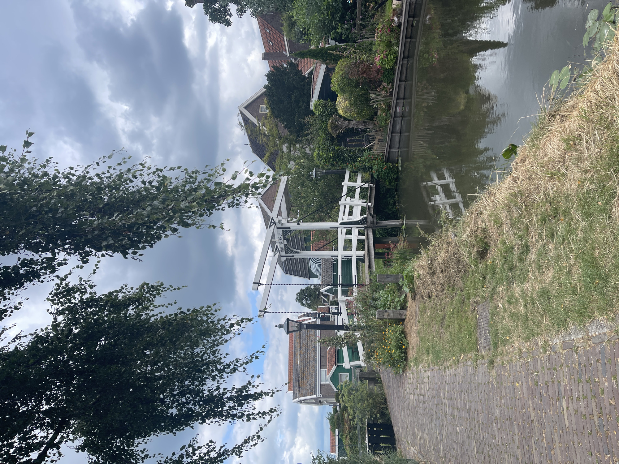 The image depicts a canal in Marken, Netherlands, with a drawbridge in the center. The drawbridge is white with a black lamp post in front of it. The canal is flanked by a brick pathway on the left and lush vegetation on the right, including trees and flowers. Several houses with red-tiled roofs and green walls are visible in the background. The sky is overcast with dramatic clouds. The scene appears tranquil and picturesque, typical of a Dutch village.