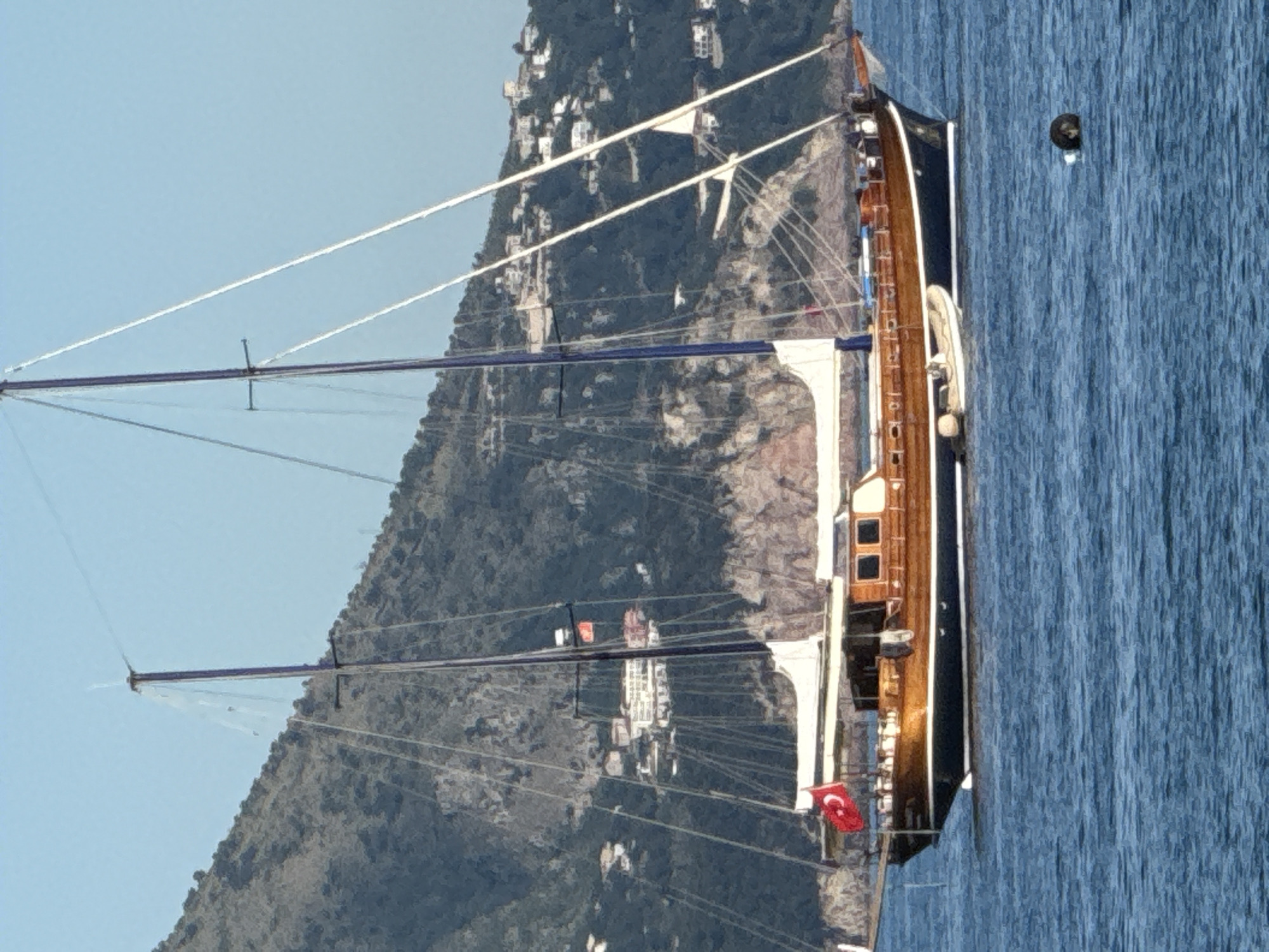 A two-masted motorsailer, characterized by its dark blue hull with a white stripe and varnished wooden superstructure, is afloat on clear blue water near Budva, Montenegro. The vessel features two prominent dark-colored masts equipped with multiple spreaders, stays, and shrouds. Off-white sails are furled along the booms and forestays. A Turkish flag (red with a white star and crescent) is prominently displayed on the bow staff, and a smaller red flag is visible higher on the main mast. A white inflatable dinghy with an outboard motor is tethered alongside the motorsailer's starboard side. Several indistinct human figures are discernible on the aft deck of the main vessel. The background consists of a steep, densely vegetated mountain slope, upon which numerous white and light-colored buildings are scattered, indicative of a coastal settlement. A dark, spherical buoy floats in the immediate foreground of the water. The sky is clear and light blue.