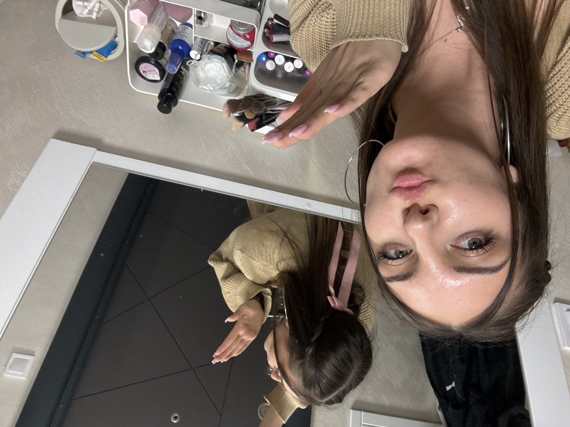 A young woman with brown hair and light skin is taking a selfie in front of a mirror. She is wearing a beige sweater and large silver hoop earrings. Her hair is styled with a pink ribbon. Her right hand is raised, with manicured nails visible. The mirror reflects the woman and the room behind her, including a dark door. On a table to the left of the mirror is a white organizer filled with various cosmetics and makeup brushes. The scene is indoors, likely in a domestic setting. There are no details in the image to indicate a specific location in Sofia, Bulgaria.