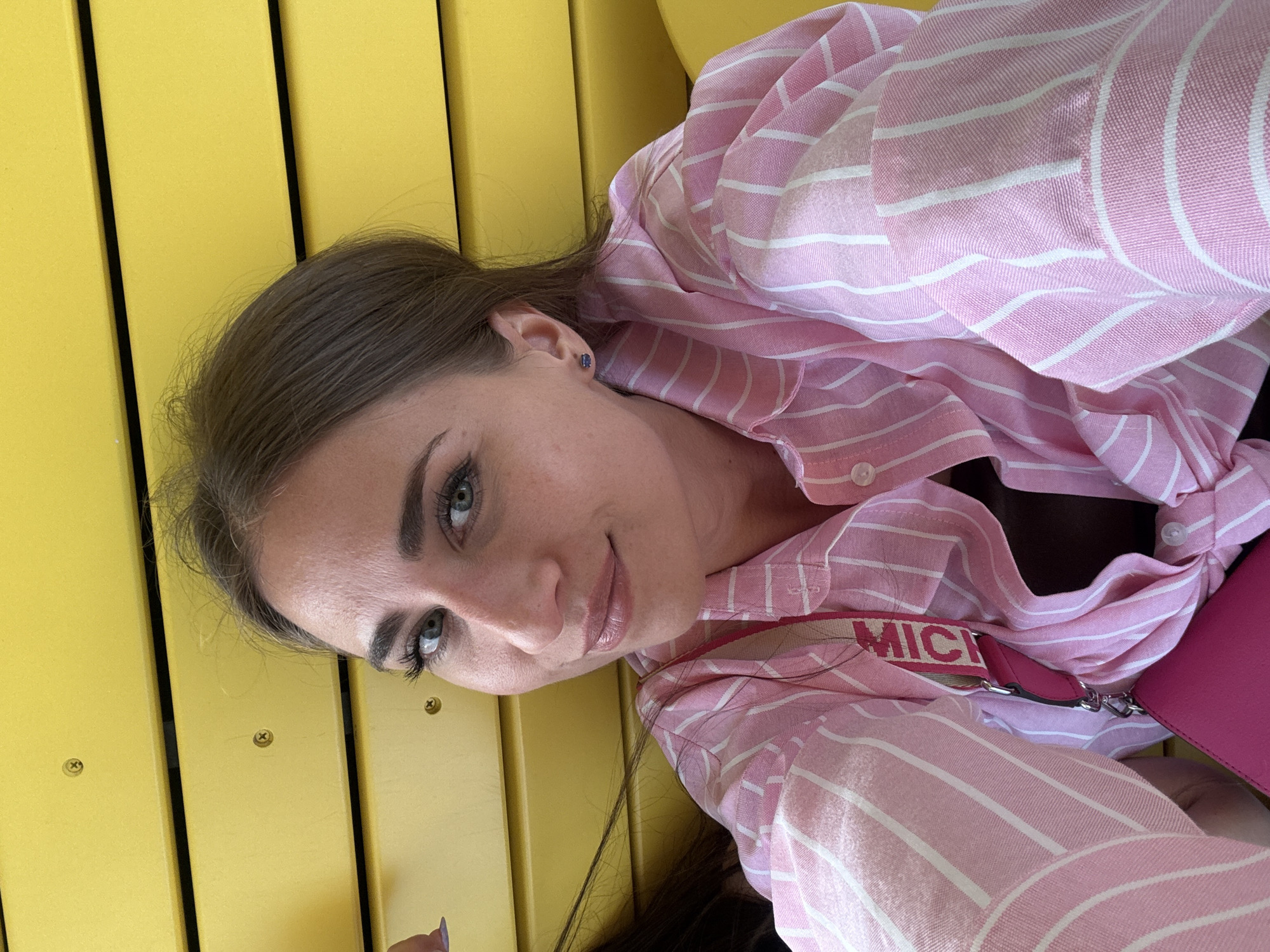 A woman with light brown hair pulled back is shown in a close-up, selfie-style shot. She is wearing a pink and white striped button-down shirt, partially unbuttoned. A pink purse strap with the word "MIC..." visible is draped across her chest. She has on small blue stud earrings. The background is a bright yellow, textured wall, possibly made of horizontal slats. The lighting is natural and appears to be daylight. The image does not provide any specific details to determine its location in Saint Petersburg, Russia.