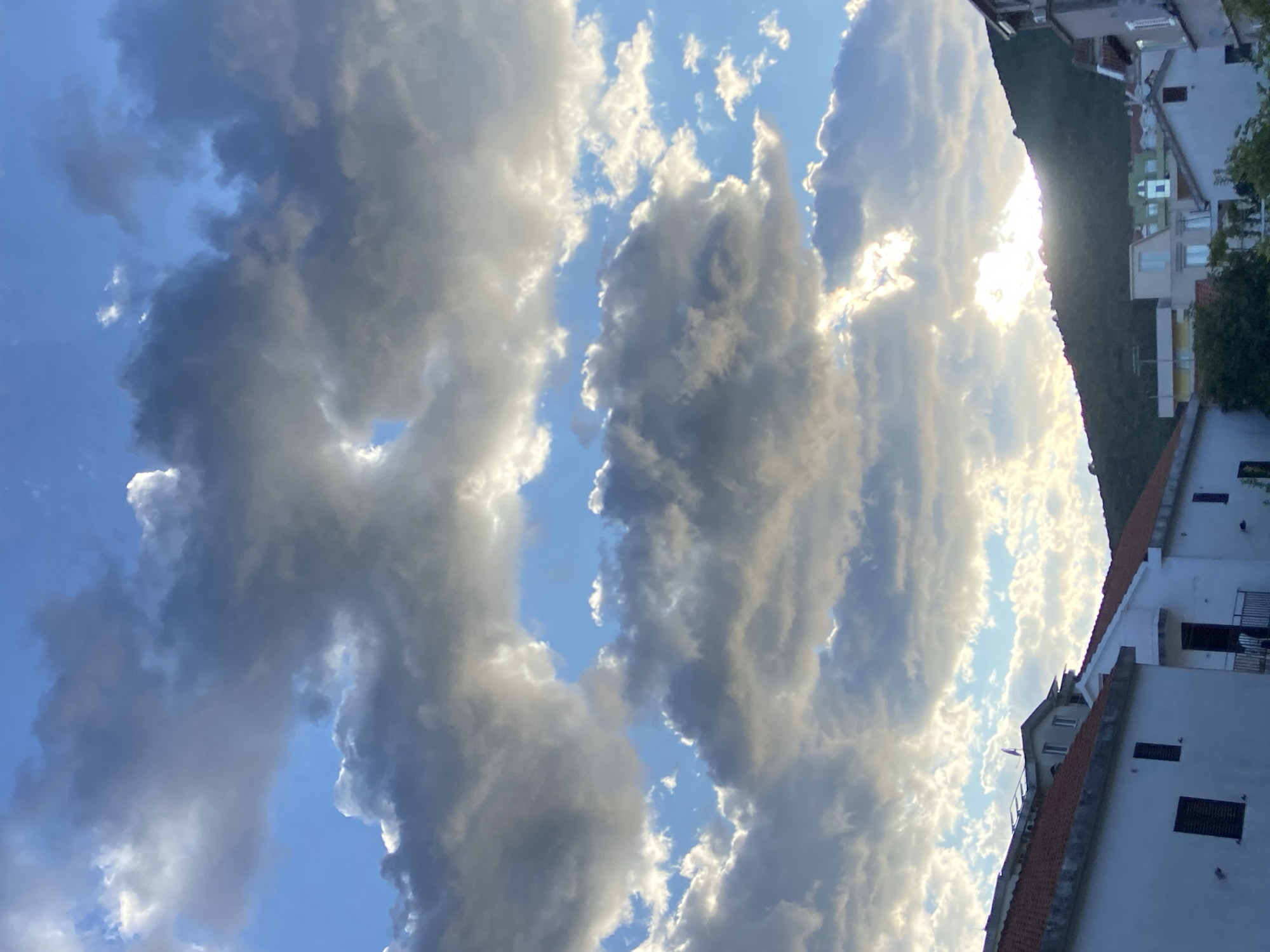 The image depicts a view of buildings in Budva, Montenegro, under a dramatic sky filled with clouds. The foreground features the rooftops and upper sections of white buildings with red tile roofs, characteristic of Mediterranean architecture. The sky above is a mix of bright blue patches and large, heavy clouds that are lit from behind by the sun, creating a luminous effect. A silhouetted green mountain range forms the backdrop on the right side of the image, with the sun setting behind it, casting a warm glow through the clouds. The overall scene suggests either a late afternoon or early evening, with the sun's rays piercing through the overcast conditions.