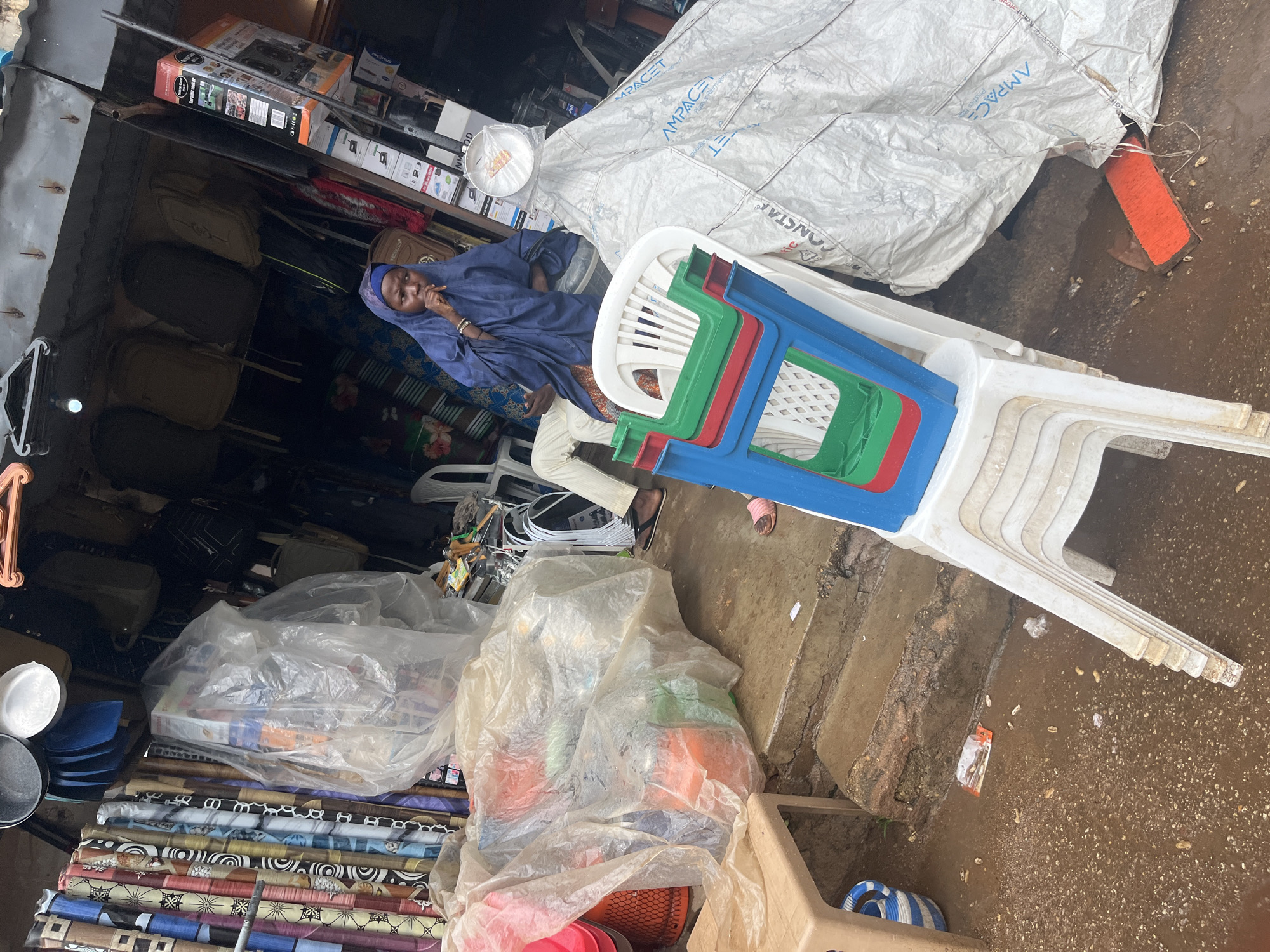 The image captures a street scene in Jos, Nigeria, likely in a market or commercial area. In the foreground, a stack of colorful plastic chairs, predominantly white, red, and blue, is visible. A person wearing a blue hijab and a white garment is seated on a white plastic chair, partially obscured by the stack of chairs. The person appears to be looking downwards with their hand near their mouth. To the left, a beige plastic table with a blue and white striped container is present. In the background, a stall displays various items, including rolls of patterned fabric and what appear to be backpacks. A large, white tarp covers some merchandise. The ground is wet and appears to be a mixture of mud and debris, suggesting recent rain or a generally damp environment. The overall impression is of a busy, open-air marketplace.