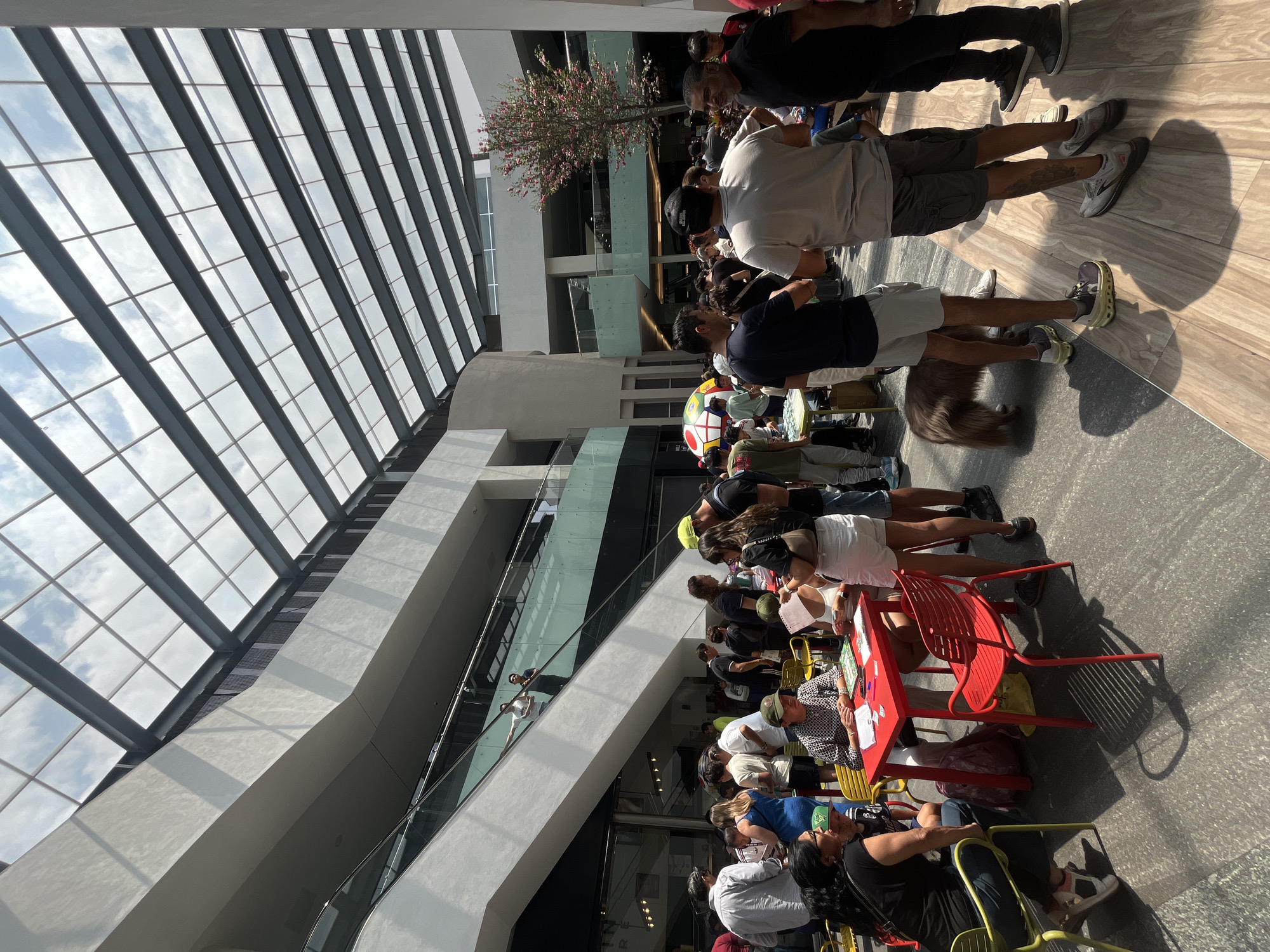 The image depicts a large indoor space, possibly a mall or atrium, in the city of Unknown, Unknown, given the presence of a high, glass-paneled ceiling allowing natural light. The floor is tiled and reflective. Numerous people are present, some standing and others seated at rows of colorful chairs arranged around small tables. Activities at the tables seem to involve writing or paperwork, implying a registration or information event. Individuals vary in age, clothing, and posture. Overhead, there is a building design with glass railings and walkways, suggesting multiple levels. A small tree is present as well.