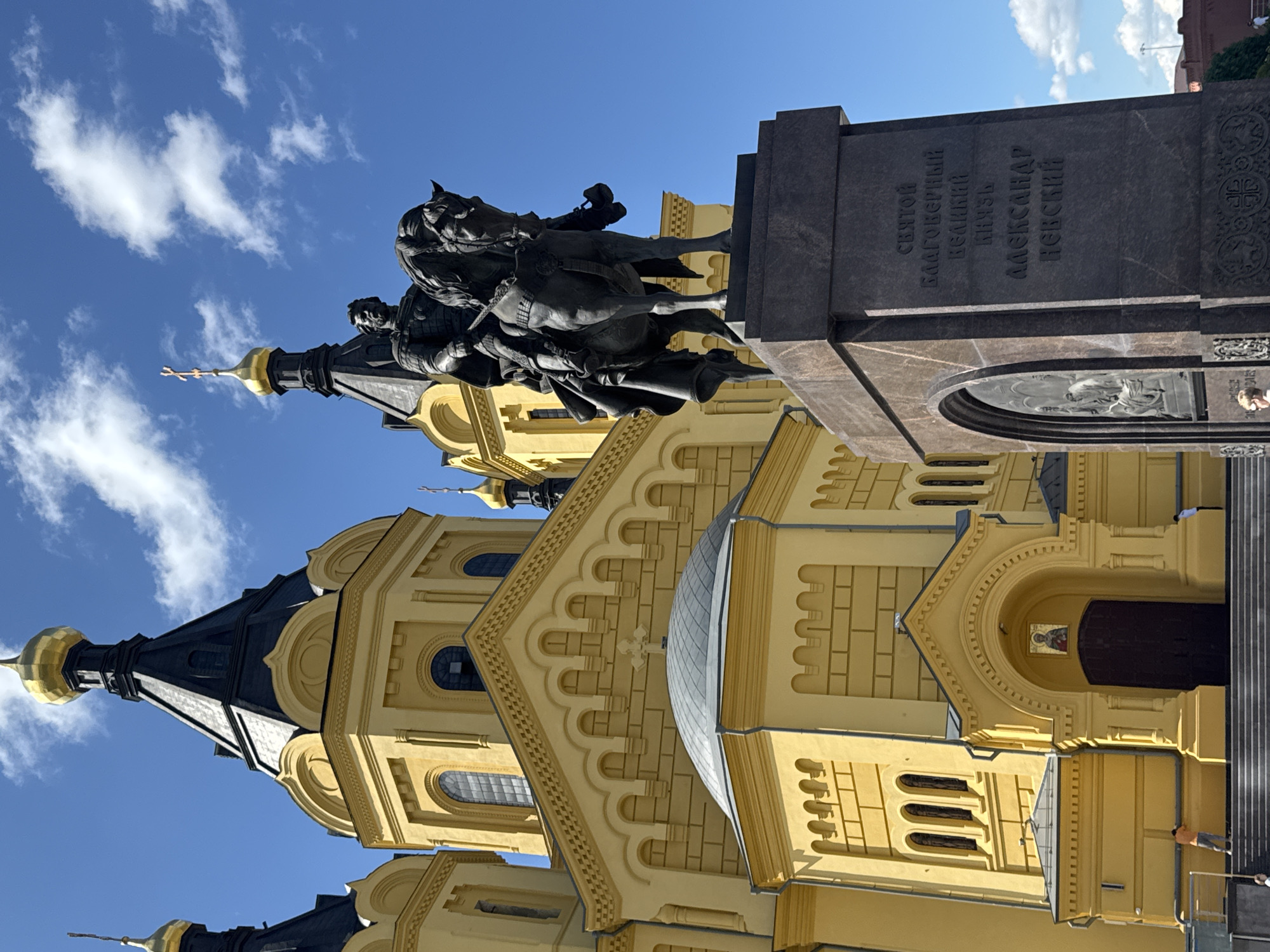 The image depicts the Alexander Nevsky Cathedral in Nizhny Novgorod, Russia. In the foreground, a bronze equestrian statue of Alexander Nevsky stands on a granite pedestal. The cathedral, painted a vibrant yellow, serves as the backdrop, with its black domes and golden crosses reaching towards a bright blue sky dotted with white clouds. The cathedral features arched windows and decorative architectural elements. The statue is positioned slightly to the right of the cathedral's entrance. The inscription on the pedestal reads "Святой Благоверный Великий Князь Александр Невский," which translates to "Saint Blessed Grand Prince Alexander Nevsky." The overall scene captures the grandeur of the cathedral and the monument on a clear day.