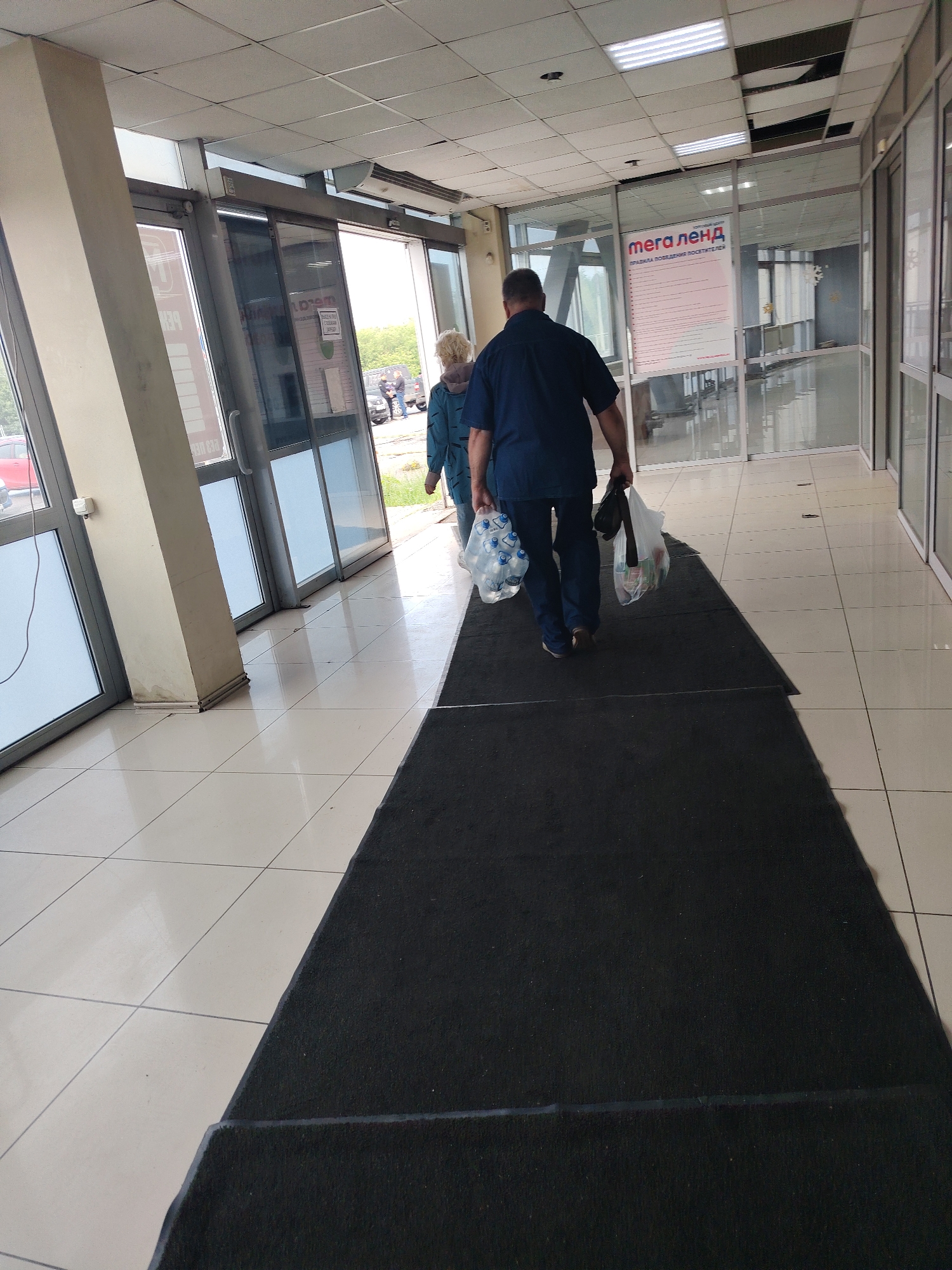 Two individuals walk away from the camera down a tiled hallway with a black carpet runner. The person in front, a man in a dark blue shirt and jeans, carries two plastic bags, one with bottles of water and the other with groceries. The person behind him, a woman in a blue jacket, also carries a bag of groceries. They are walking towards automatic glass doors that lead outside, where a red car is visible. A sign for "Мега Ленд" is posted on a glass partition to the right. The hallway is well-lit with fluorescent ceiling lights, and there is a large pillar on the left side. The floor is tiled with large white squares.