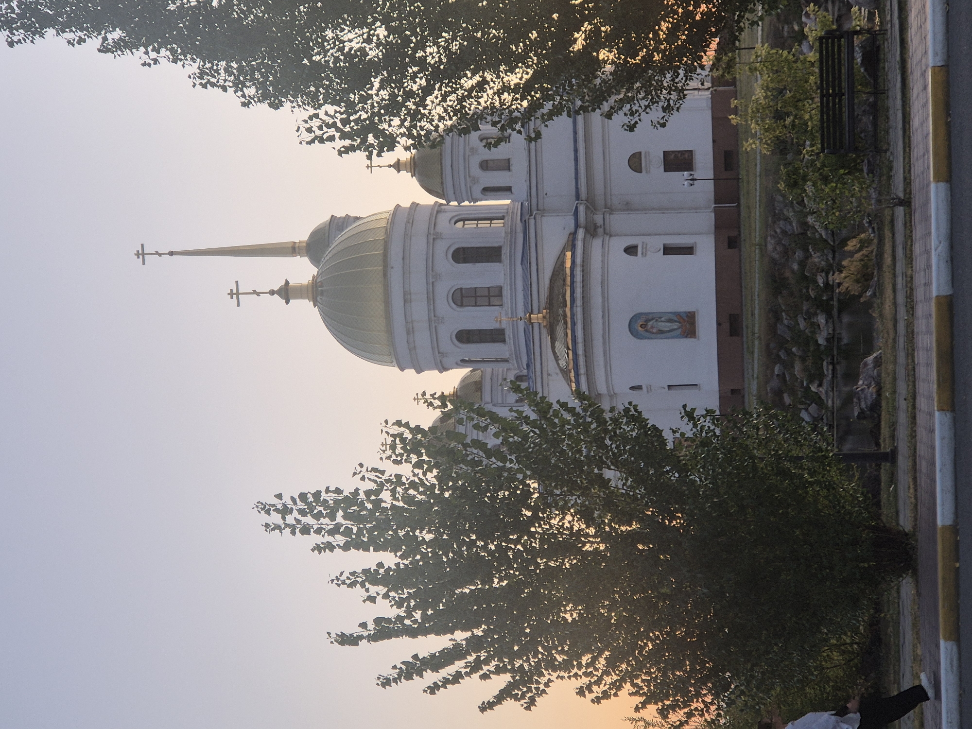 The image depicts a white Orthodox church with golden domes, likely the Church of the Intercession of the Mother of God in Kokshetau, Kazakhstan, seen from a slightly low angle. Two prominent trees, one on the left and one on the right, partially obscure the view of the church. The sky is a gradient of pale blue and soft orange, suggesting either sunrise or sunset.
In the foreground, a person in a white shirt and dark pants is walking from left to right across a paved walkway. A park bench is visible on the right side of the image, situated near a rocky area with a body of water. The church itself has arched windows and a fresco of a religious figure on its facade. A lamppost stands near the church. The overall scene conveys a sense of tranquility, bathed in the warm light of dawn or dusk.