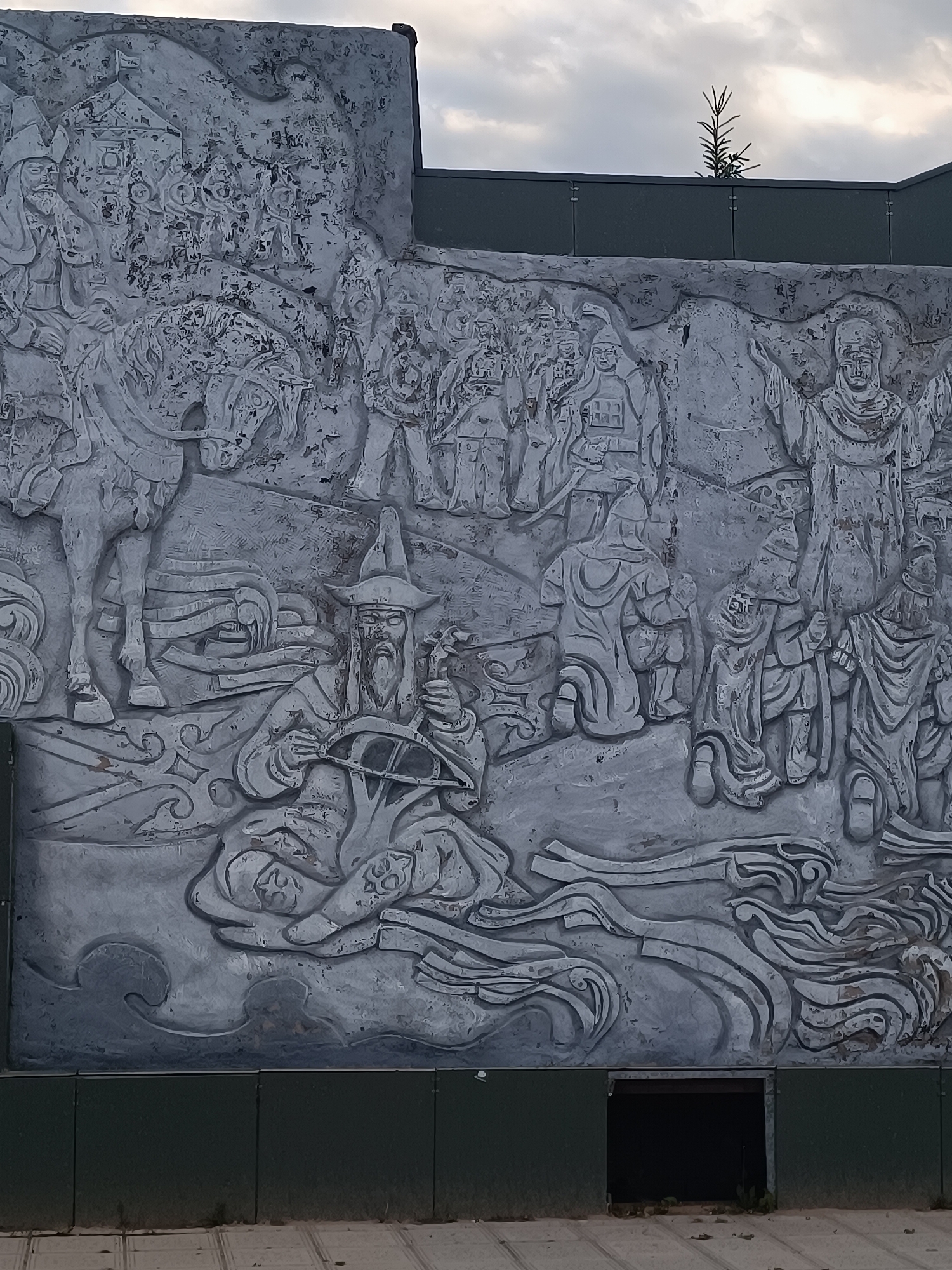 The image shows a large, gray bas-relief mural on a building in Kokshetau, Kazakhstan. The mural depicts a variety of figures, including men on horseback, seated figures with elaborate hats, and standing figures in robes. The style is reminiscent of traditional Central Asian art. Below the mural is a dark green panel, and at the bottom of the panel is a square opening leading into the building. Above the mural, a green fence and a small evergreen tree are seen against a cloudy sky. The ground in front of the mural is paved.