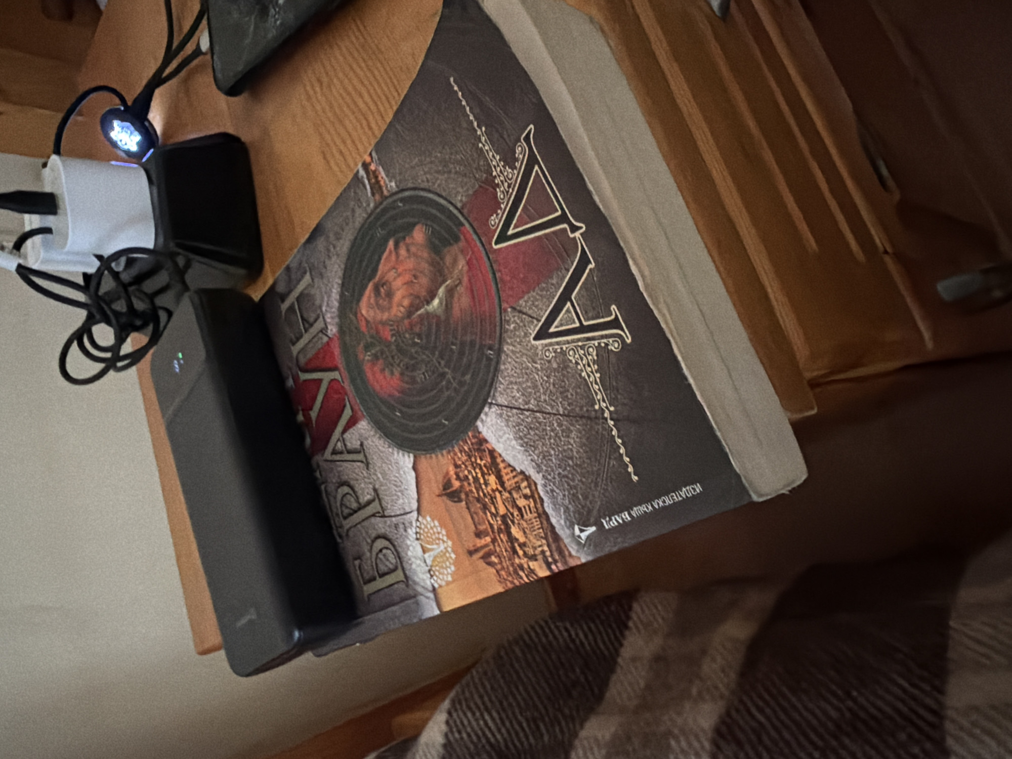 A close-up, angled shot shows a book with a dark cover displaying images of a city and a man's face. The title appears to be in Cyrillic. On top of the book is a black power bank. To the right of the book, two white USB wall adapters are plugged into an electrical outlet, with one also connected to a small, illuminated blue device, and a black cable extending from it. A part of a wooden drawer is visible at the bottom. The overall scene is set on a wooden surface, likely a nightstand. The location in Veliko Tarnovo, Bulgaria cannot be determined from the image.