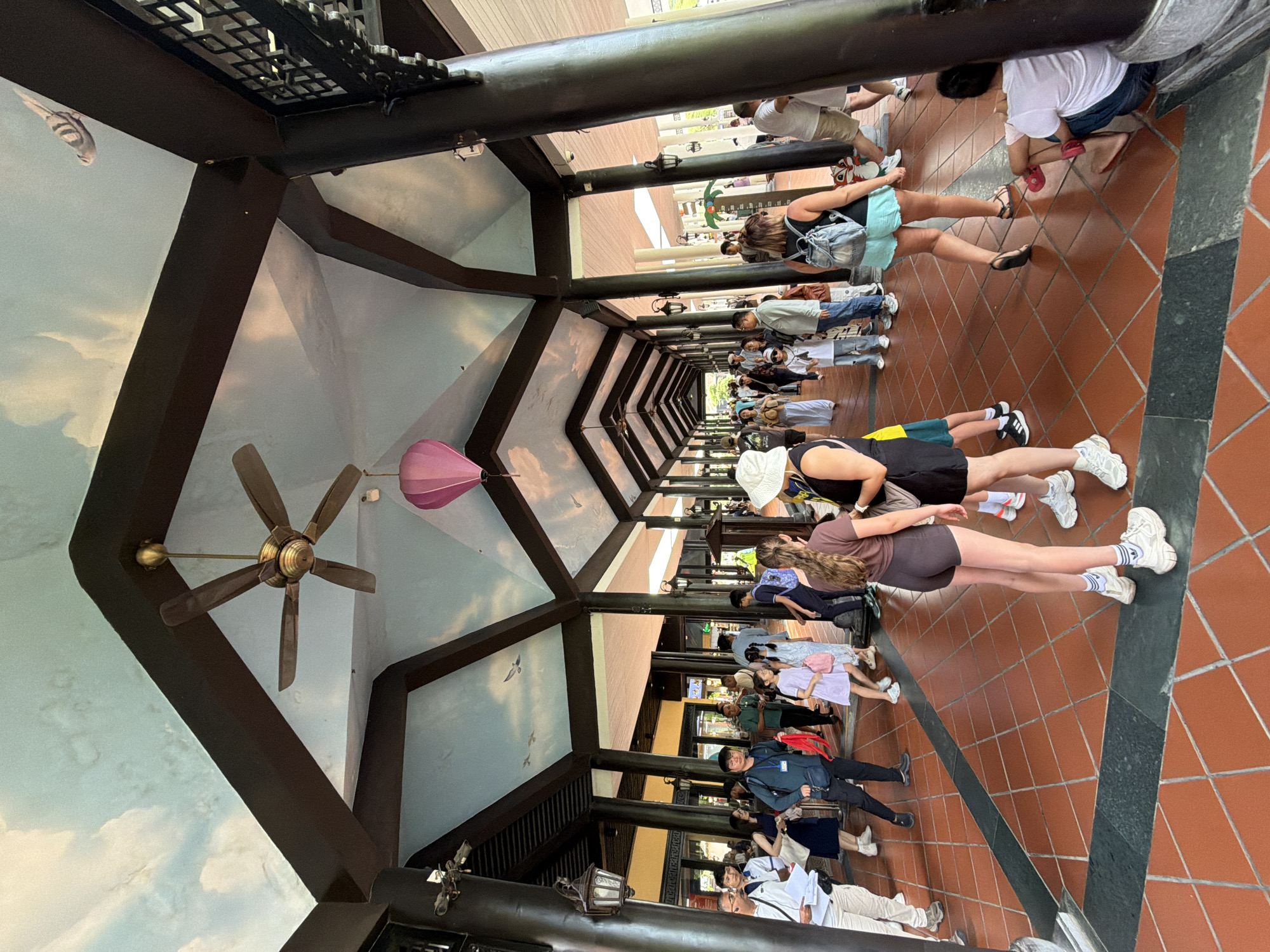 The image depicts an outdoor covered walkway, likely part of a tourist attraction or transit point in Đà Nẵng, Vietnam. The walkway features a high, pitched ceiling adorned with a sky mural featuring clouds and birds, and supported by dark wooden pillars. Several ceiling fans are visible, and a pink hot air balloon-shaped lantern hangs from the ceiling. The floor is tiled with reddish-brown square tiles arranged in a diamond pattern, bordered by dark grey bands.
A diverse group of people, appearing to be tourists, are walking through the covered area. They are dressed in casual summer clothing, including shorts, t-shirts, dresses, and hats. Some individuals are carrying backpacks or bags. The people are in motion, some in conversation or looking around.
Notable individuals include a woman in the foreground wearing a white hat and black top, and another woman next to her wearing a brown top and dark shorts. Further down the walkway, a man in a blue shirt and dark pants is prominent.
The environment suggests a warm climate, given the attire of the people and the presence of ceiling fans. The architecture and the sky mural contribute to a potentially whimsical or themed atmosphere. The scene captures a moment of pedestrian traffic and leisure activity within a public space.