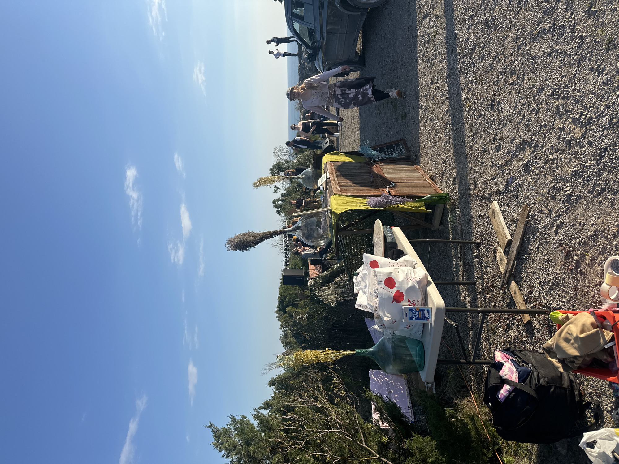 The image depicts an outdoor scene in Bigova, Montenegro. The foreground features a white plastic table loaded with items, including large glass bottles, bags with red poppy designs, a black backpack, and other miscellaneous objects. Behind the table is a rustic wooden structure, possibly a stall or makeshift shop. Several people are present, some seated and others standing near the structure. A woman walks in the background, alongside a dark-colored car parked on a gravelly surface. In the distance, the coastline is visible, separating the sky from the land. The sky is blue with scattered white clouds. Green trees surround the area.
