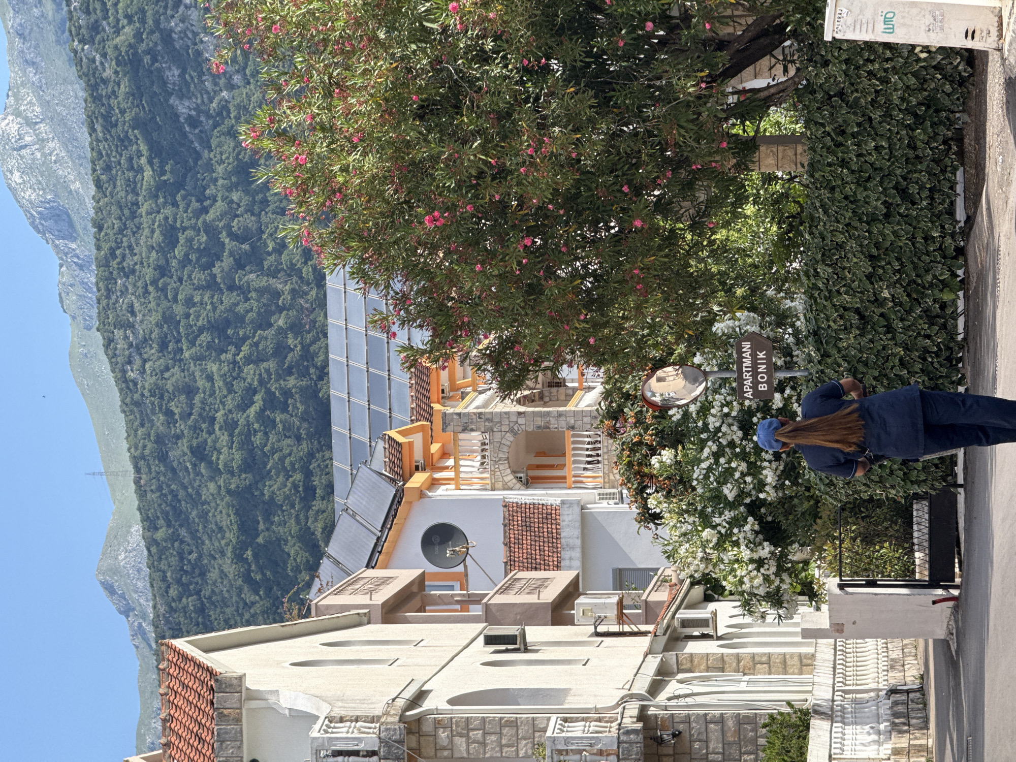 The image shows a street scene in Budva, Montenegro, with a view of a building with solar panels on the roof and mountains in the background. A person wearing a blue outfit and a blue hat is walking on the sidewalk. The building has a sign that reads "APARTMANI BONIK". There are various plants and greenery alongside the building and the sidewalk. The architecture appears typical of the region, with light-colored stone and tiled roofs.