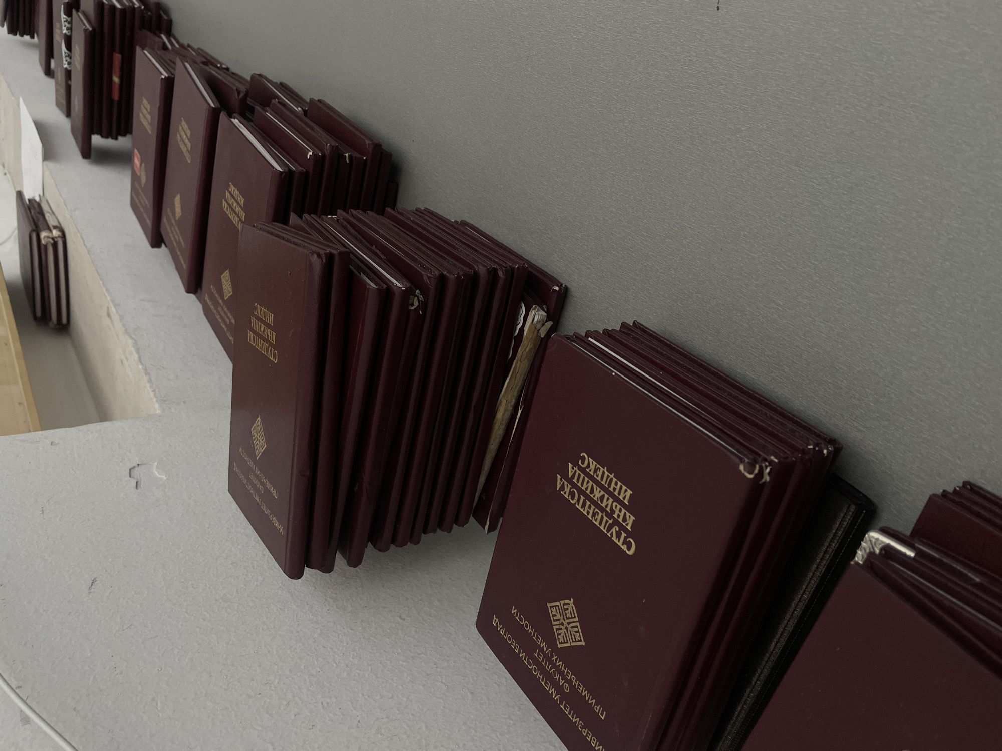 The image displays numerous dark red, hardbound books, identified as "СТУДЕНТСКА КЊИЖИЦА ИНДЕКС" (Student Book Index), arranged in multiple rows on a light grey surface and leaning against a plain grey wall. The gold Cyrillic text on the covers indicates they belong to the "УНИВЕРЗИТЕТ УМЕТНОСТИ БЕОГРАД" (University of Arts Belgrade) and specifically the "ФАКУЛТЕТ ПРИМЕЊЕНИХ УМЕТНОСТИ" (Faculty of Applied Arts).
The scene is an indoor setting, likely an administrative office or a collection point within the university in Belgrade, Serbia. The books appear to be stored, awaiting distribution, or organized for administrative tasks, such as student registration or record keeping. Some of the books show visible signs of wear, including minor scuffs, frayed edges, and slightly bent corners, suggesting they have been frequently handled. The background is a solid grey wall, and the surface the books rest on is a textured light grey. The overall impression is that of an organized collection of official student academic records.