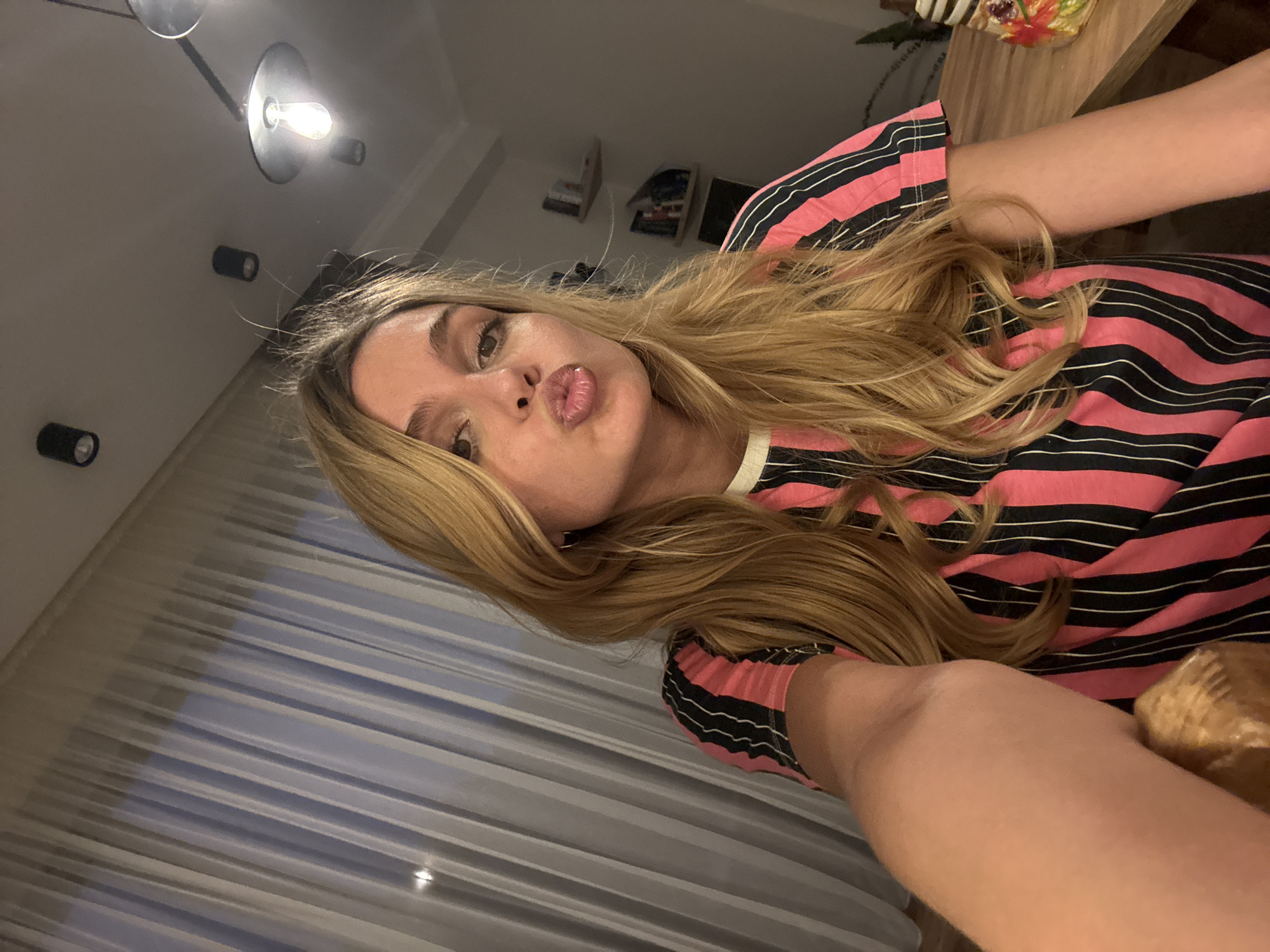 An indoor selfie captures a young woman with long, flowing blonde hair, fair skin, and prominent eyebrows. She is making a "kissing" or "pouting" face towards the camera. She wears a striking pink and black striped long-sleeved top.
The setting appears to be a modern room, likely a home, during the evening or night, indicated by the use of artificial lighting. The ceiling features several light fixtures, including recessed spotlights and a decorative fixture with an exposed bulb. In the background, there are glimpses of shelves or wall decor, and a wooden surface, possibly a table or cabinet, with a small, colorful decorative item on it. To the left and below the woman, a white pleated curtain or blind covers a window. Her arm is visible, along with glimpses of her leg/knee area and what might be a furry pet or blanket to the far right. The overall mood is casual and playful. There is no visible text in the image.