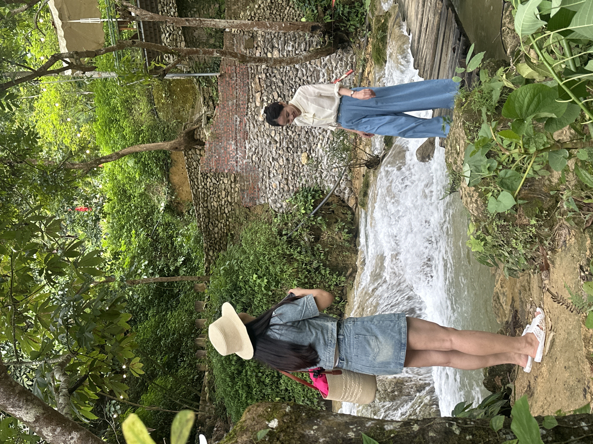 Two women are standing in a natural environment with a flowing stream and lush greenery. The woman on the left is wearing a straw hat, a denim jacket, and a denim skirt, and is holding a straw bag with a pink interior. She is standing on a rocky, earthy area near the stream. The woman on the right is wearing a white blouse with a bow tie and blue wide-leg pants. She is walking across a wooden bridge that spans the stream. The background features a stone and brick wall, trees, and dense vegetation, suggesting a rural or natural setting. The overall scene depicts a relaxed outdoor activity in a picturesque landscape. While the provided location is Cổ Lũng Commune, Vietnam, the image itself does not contain explicit visual cues to confirm this specific location.