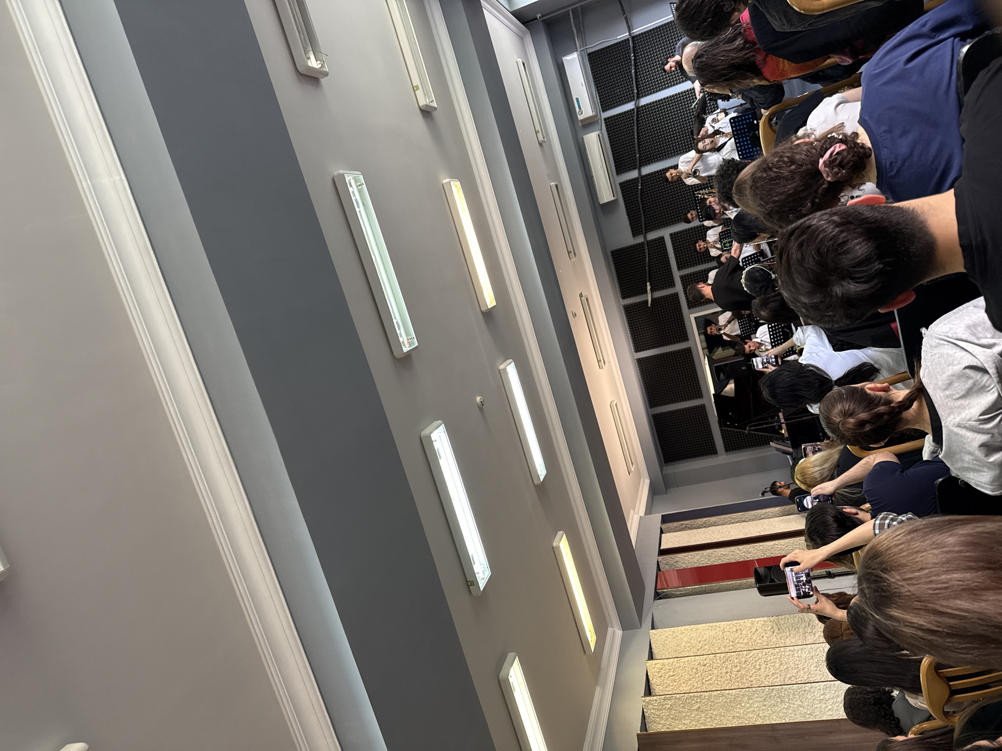 The image shows a group of people sitting in an auditorium or performance space, seemingly in Yerevan, Armenia. The space appears to be modern, with a gray and white color scheme and fluorescent lights on the wall. The people are seated in rows on tiered levels, facing a stage where musicians are performing. Many audience members are using their phones to take pictures or videos of the performance. The walls are lined with soundproofing panels.