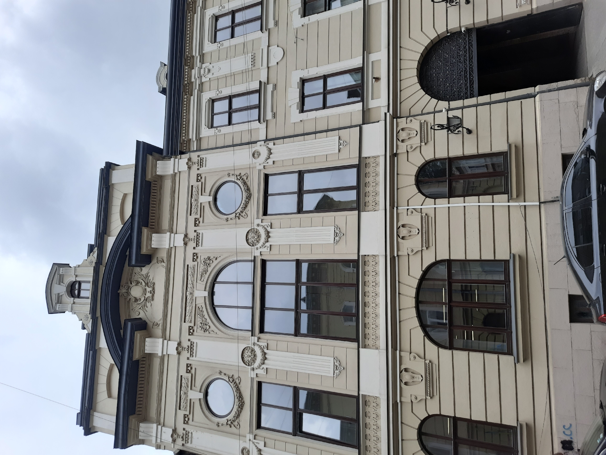 The image shows a light beige building with intricate architectural details and multiple windows, located on a street in Lviv, Ukraine. The building features a mansard roof with a central dormer window and decorative elements above the windows on the upper floors. The windows are large and dark-framed, with some reflecting the cloudy sky. The facade is adorned with decorative moldings, reliefs, and pilasters. On the ground floor, there is a large arched entrance with ornate ironwork. A black car is parked in front of the building, partially obscuring the lower part of the facade and a street lamp. The sky is overcast, indicating a cloudy day.
