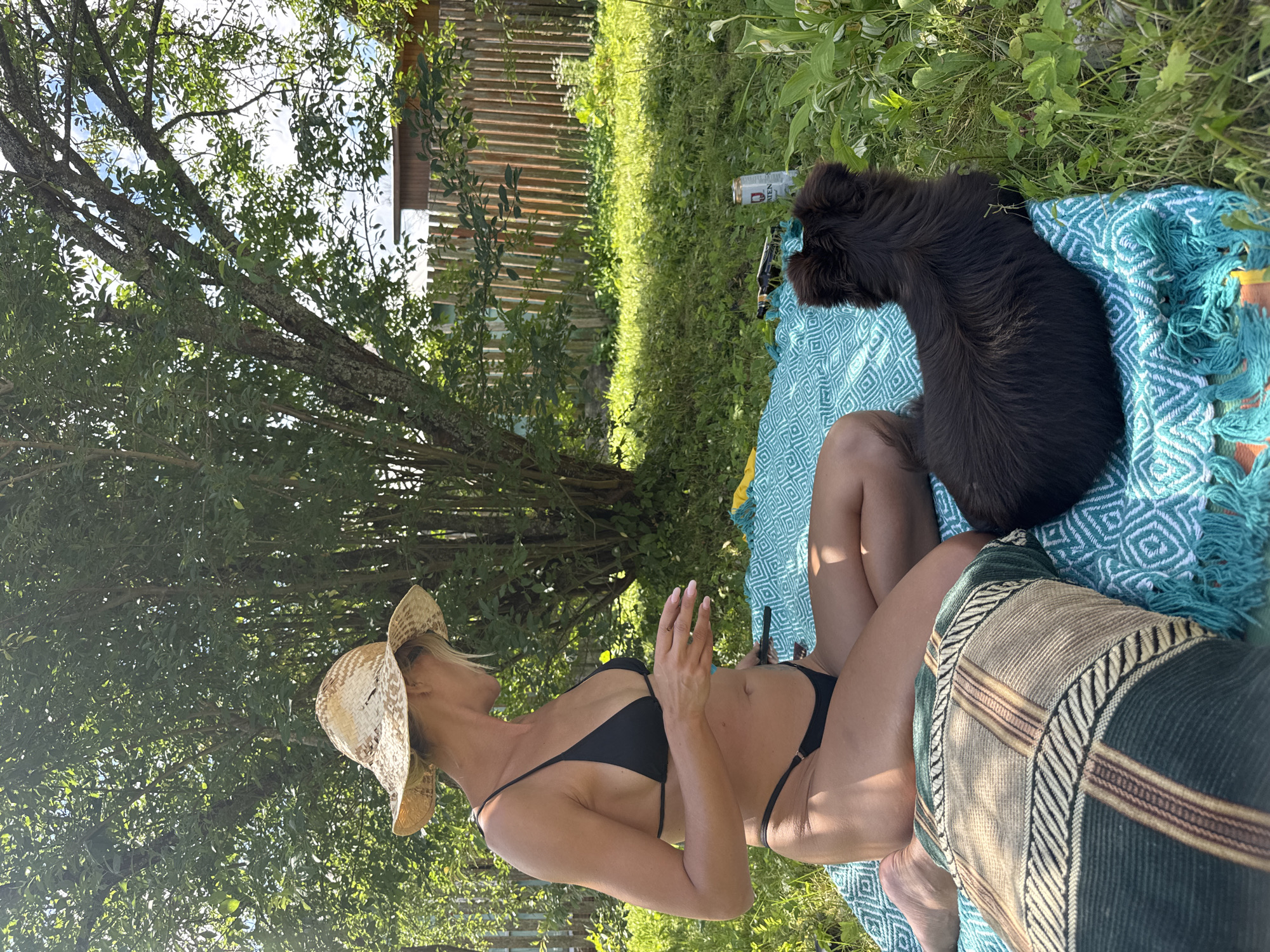 A blonde woman wearing a straw hat and a black bikini is sitting on a teal blanket in a grassy backyard. She is looking to her left, with her right hand held up, as if holding something. A small, dark brown dog is lying on the blanket to her right, facing away from the camera. In the background, a wooden fence runs along the edge of the property, with trees and foliage visible behind it. A tall can of beverage and a pair of sunglasses are also on the blanket near the dog. The scene appears to be in a suburban or rural setting, possibly a dacha or backyard in a village.