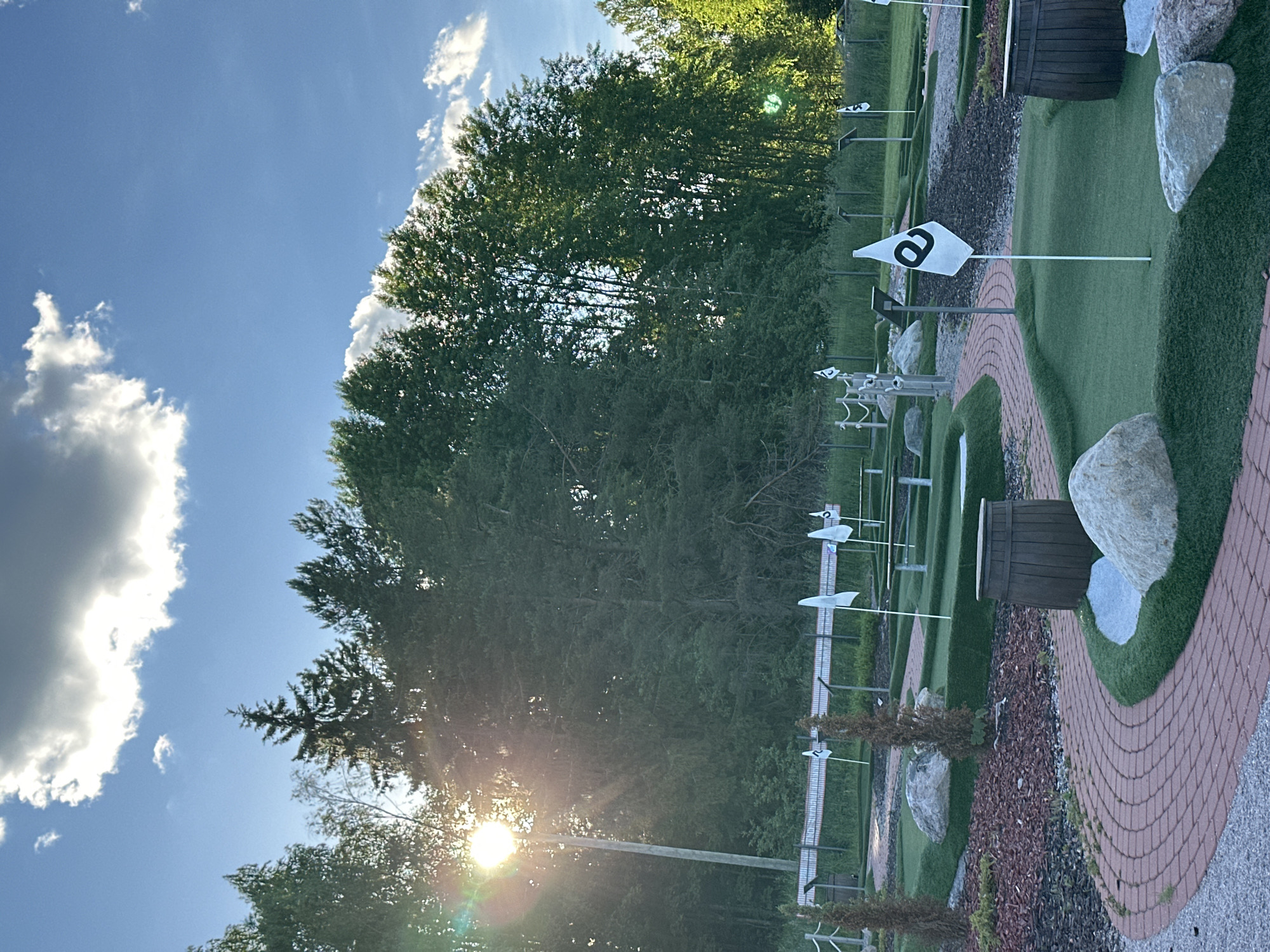 A sunny, late afternoon scene at a mini-golf course depicts a winding red brick path curving through artificial green turf. Several white flags with black logos, resembling the letter "e," are planted on the course, marking holes. Large grey rocks are strategically placed as obstacles. Two wooden barrels, weathered and dark, sit near the path. In the background, a dense line of evergreen trees forms a natural backdrop. A bright sunbeam flares from the upper left of the image, partially obscured by clouds. The sky is blue with scattered white clouds. The location appears to be outdoors, likely in a park or recreational area, with no people visible in the frame.