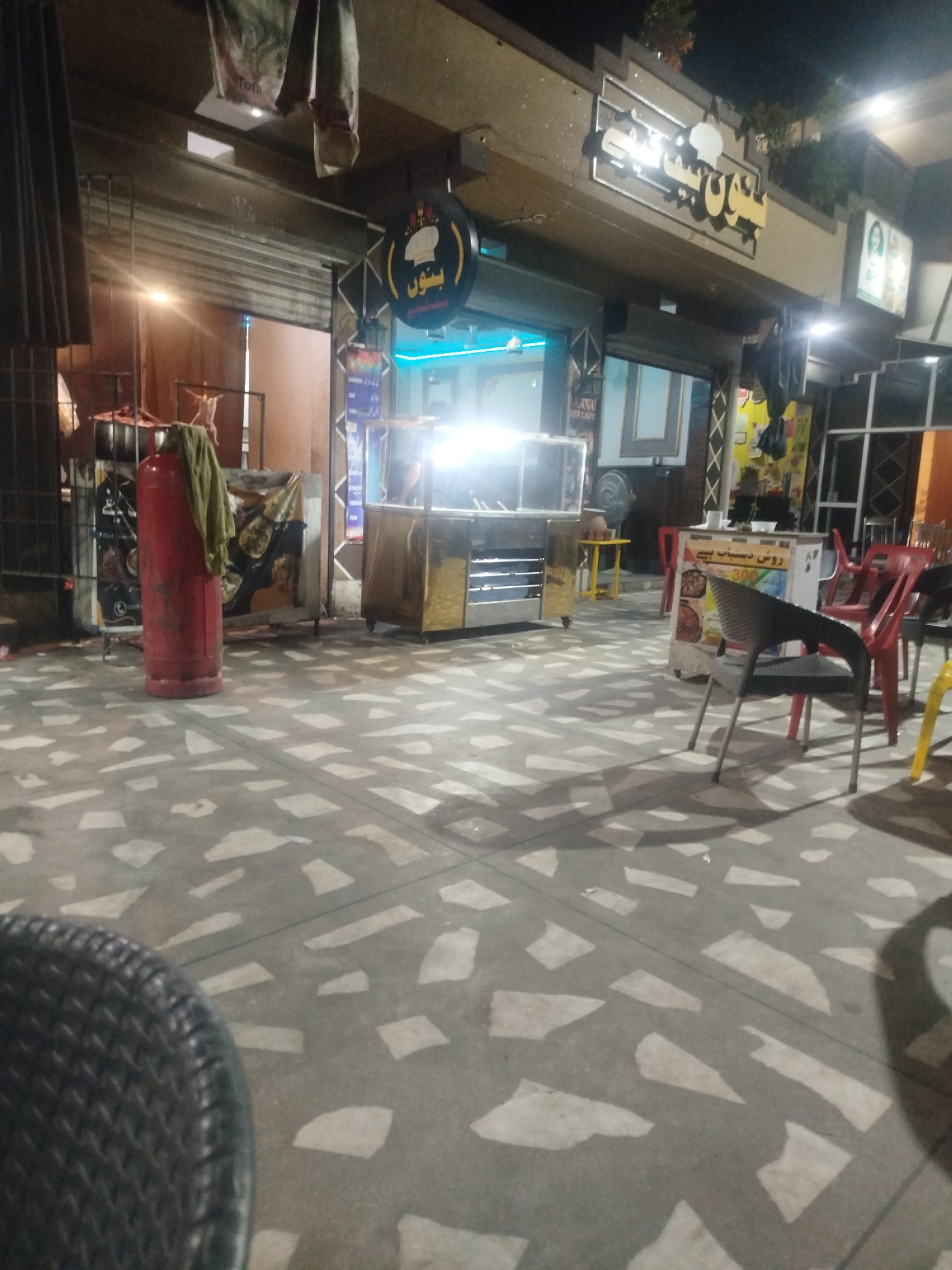 The image shows a street-level view of a restaurant or food stall establishment in Peshawar City Tehsil, Pakistan at night. The scene is illuminated by artificial lighting, casting shadows on the patterned concrete ground.
In the foreground, the textured seat of a dark chair is visible on the left. The middle ground features a red gas cylinder with a green cloth draped over it, positioned near a food stall with a rolled-up shutter. Another food stall with a stainless steel counter and overhead lighting is present further back. Various chairs, including red and black plastic ones, are arranged around small tables. In the background, a building with a prominent sign featuring Arabic script and a chef's hat emblem is visible. Another sign with images of food is also present. The overall environment suggests an outdoor dining or take-away area. There are no clear individuals visible engaging in specific actions within the frame.
