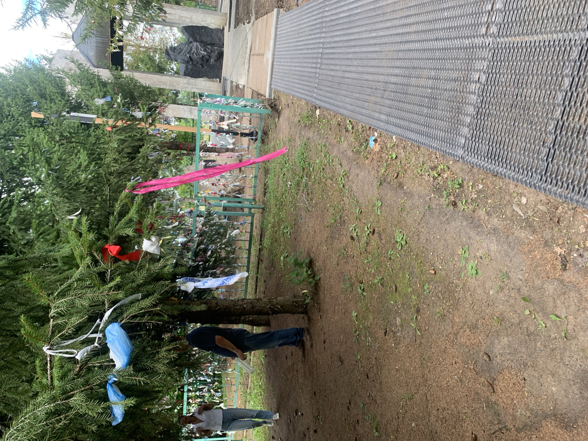 In the city of Arbuzov-Baran, Russia, a scene unfolds around a natural memorial adorned with prayer ribbons tied to trees and a fence. Two individuals are present: a woman in a white shirt and blue jeans walking away from the camera, and a man in a dark blue shirt and jeans standing near a tree, holding a clear plastic bottle. The environment is a wooded area, with lush green fir trees dominating the foreground. The ground is covered in a mixture of dirt, pine needles, and small green plants. Behind the trees, a teal-colored metal fence is visible, also decorated with numerous colorful ribbons. Further in the background, a memorial structure is partially obscured by the trees. It appears to be made of stone pillars and a dark, abstract sculpture. A metal walkway extends from the right side of the frame towards the memorial area.