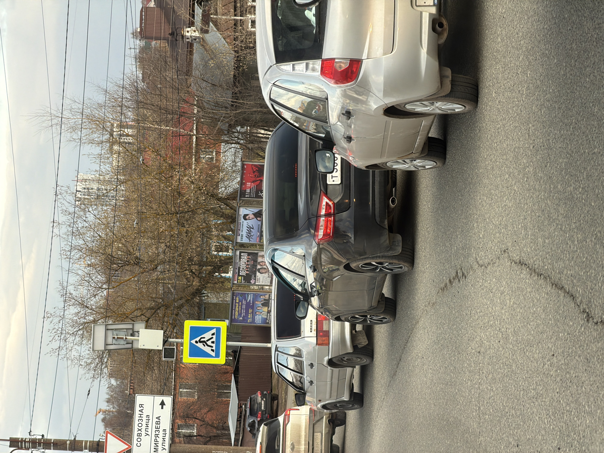 The image depicts a street scene in Izhevsk, Russia, on an overcast day, likely during late winter or early spring, given the bare branches of the trees. The view is from the side of the road, looking along a line of parked cars.
In the foreground, several vehicles are visible, including a silver hatchback, a dark sedan, and a lighter-colored car bearing a "LADA" badge. The asphalt road surface shows some wear and markings. A prominent blue and yellow square sign indicates a pedestrian crossing. Below it, a white road sign with an arrow pointing straight ahead displays Cyrillic text: "СОВХОЗНАЯ улица" (Sovkhoz Street) and "МИРЯЗЕВА улица" (Miryazeva Street). A fragment of a car license plate shows "6BM 159".
Utility poles with numerous overhead wires run parallel to the street. In the mid-ground and background, various billboards are visible through the trees, advertising events with text such as "ЛЕСОПОВАЛ" (Lesopoval), "AKMAL", "20.21 MAR/19.00", "17.06", and "ПОДВОДНЫЙ МИР" (Underwater World). Residential buildings with red-brick facades are scattered among the trees, and a taller, more modern building can be seen in the distant skyline under the grey sky. No people are visible, but the parked cars and street signs suggest an active urban environment.