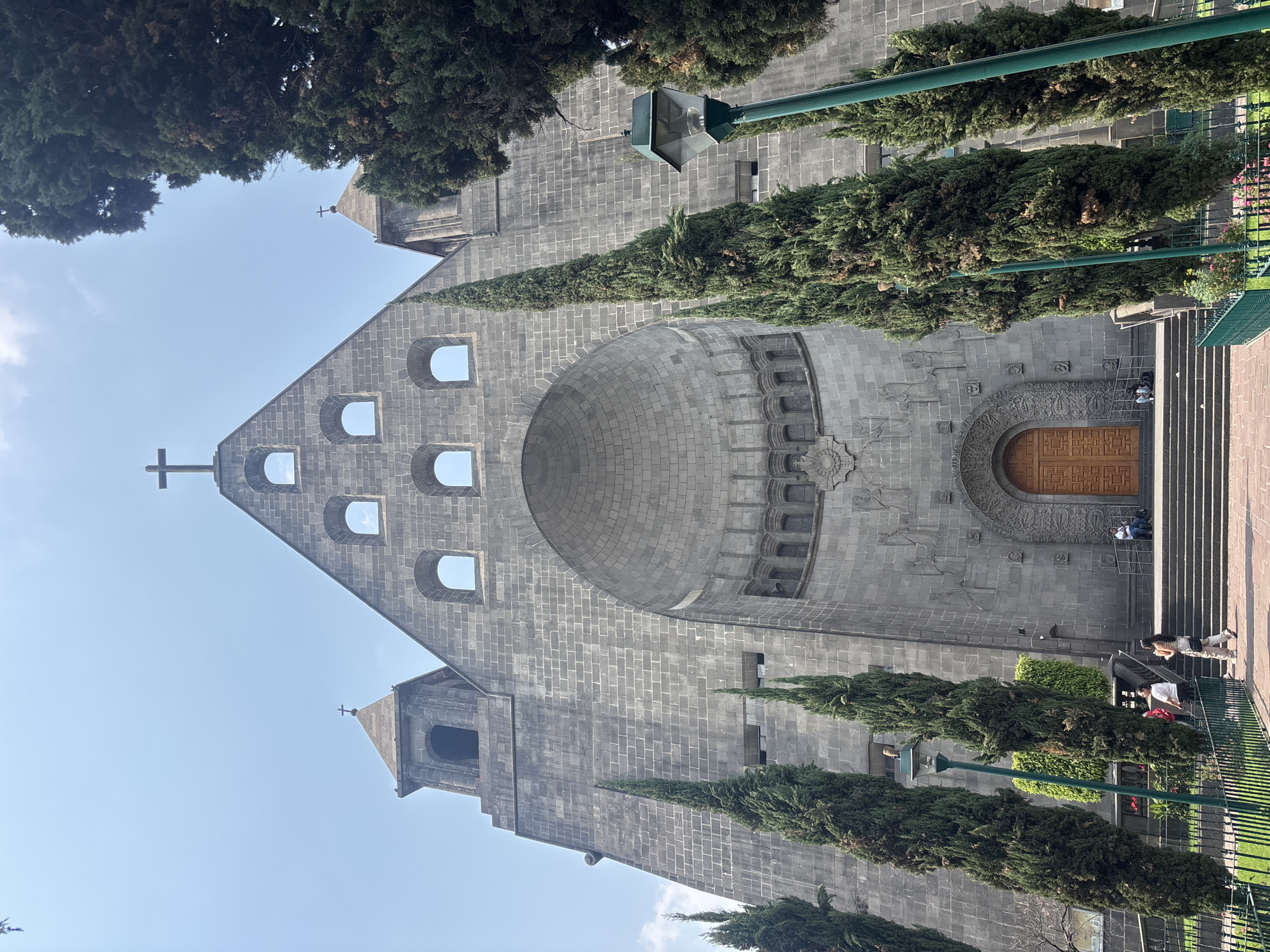 The image features the Templo Expiatorio a Cristo Rey, also known as the Antigua Basílica de Guadalupe, located in Mexico City, Mexico. The building's gray stone facade is visible, with a triangular pediment adorned with a cross, arched windows, and a semi-circular architectural detail above the main entrance. A large wooden door is centered at the base of the structure. The front of the basilica is bordered by trees and green lamp posts, and a metal fence. Steps lead up to the entrance where a few people can be seen. The sky is visible above the building, partially obscured by trees.