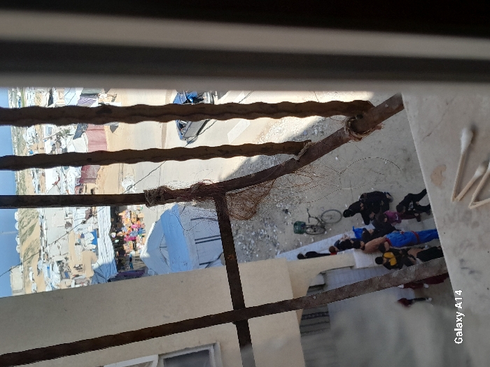 The image depicts an elevated view through a window in the city of Khan Yunis, Palestinian Territory, looking down upon a street level scene and across to an informal settlement. Three rusty, twisted vertical metal bars and a partial frame on the right segment the view. On a concrete window ledge in the immediate foreground, three wooden-handled cotton swabs are present.
Below, on a dirt path adjacent to a white multi-story building, a group of approximately 10-15 individuals, including adults and children, are gathered. Several individuals wear dark clothing, and some women appear to wear head coverings. A bicycle with a green basket on its rear rack is parked upright near this group. Further back, a light-colored sedan with its trunk open is parked on a paved street. A white tent-like structure is situated to the left of the sedan.
The background features a dense, expansive informal settlement, characterized by numerous makeshift structures constructed from corrugated metal, tarps, and other materials. Individuals are visible moving between these structures. A market stall displaying colorful goods is discernible among the buildings. In the far distance, barren hills are visible under a clear sky, with a tall, slender mast or tower situated on one of the distant elevations.