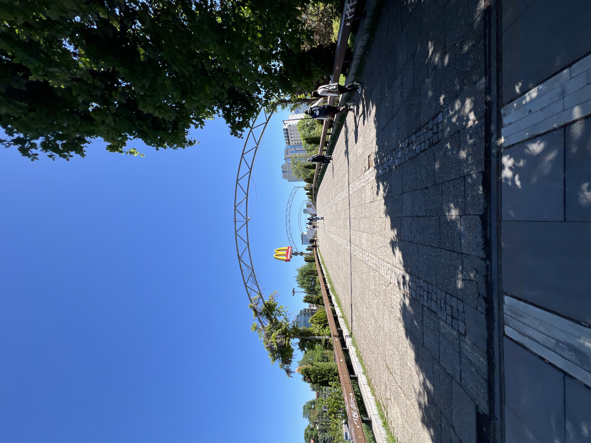 This image shows a pedestrian walkway in Sofia, Bulgaria. A large McDonald's sign is visible along the path, which is lined with trees and a metal railing. People are walking in the distance. The sky is clear and blue. The ground is paved with gray bricks, and there are patches of sunlight and shadow.
