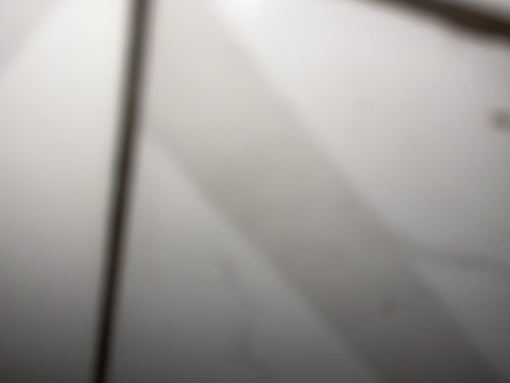 The image is a blurry, close-up shot of a white, textured surface, possibly a ceiling or wall. Shadows are cast diagonally across the surface, suggesting the presence of an object or structural element above and to the left. A dark, vertical line, potentially a shadow or a thin structural element, runs down the right side of the image. The lighting appears to be artificial and diffused. There are no discernible objects, people, or actions taking place. The location is unknown.