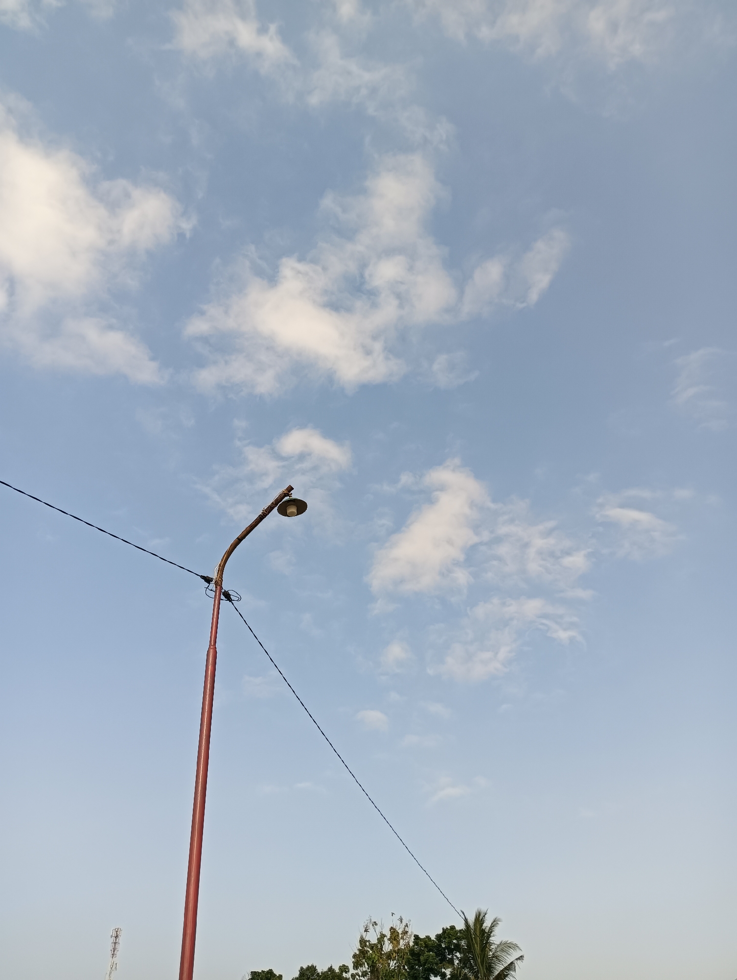 An outdoor, low-angle, vertical shot captures a red lamppost and wires against a pale blue sky dotted with white clouds. The lamppost is positioned on the left side of the frame, with its curved top supporting a light fixture that is not illuminated. Two dark wires extend horizontally from the lamppost, disappearing out of the frame. In the background, the upper portions of green trees and a distant radio tower are visible. The image appears to have been taken in Cluwak, Indonesia, though no specific landmarks are identifiable beyond the general landscape. The scene is devoid of any human presence or activity.