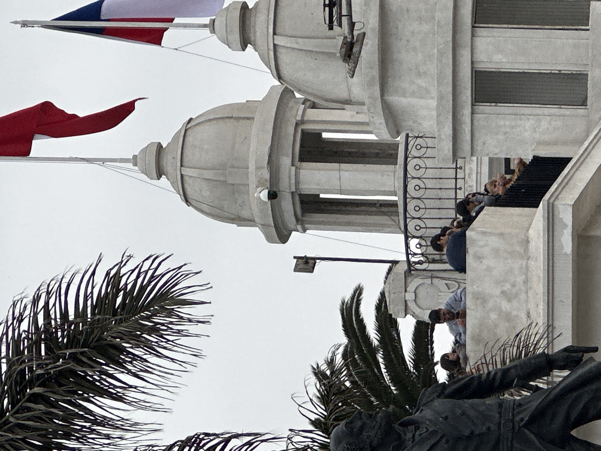 The image captures a scene in Arica, Chile, featuring a building with two prominent domes and a balcony. People are visible on the balcony, looking outwards. In the foreground, there is a statue of a man holding a gun, and palm trees are present on either side of the statue. Two flags are flying atop the building, one red and the other with blue, white, and red. The sky is overcast.