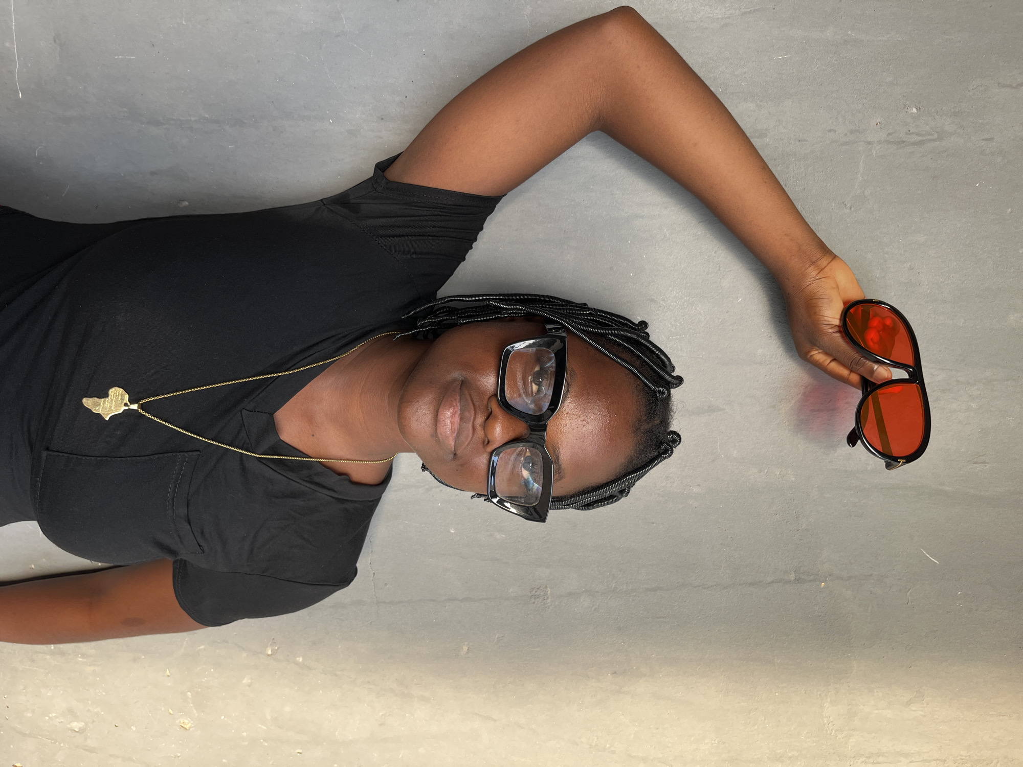 A person is lying on a grey surface, likely a floor or concrete area. The person is wearing a black t-shirt and eyeglasses. They also have on a necklace with a pendant shaped like the continent of Africa. The person's hair is styled in braids. In the person's right hand are sunglasses with red lenses. No details provide that the scene takes place in Badagry, Nigeria.