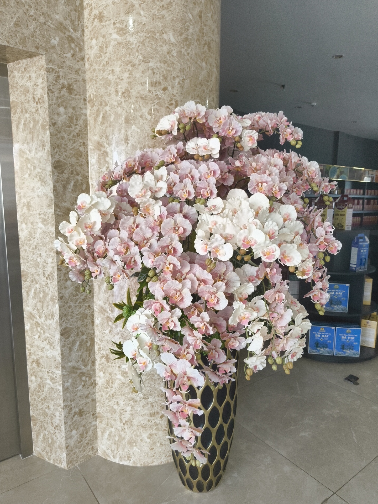 This is a full-shot, eye-level, indoor photograph of a large floral arrangement. The arrangement consists of numerous pink and white orchids with green stems and leaves, displayed in a tall, patterned black and gold vase. The vase is positioned in front of a beige, marbled wall. To the left of the floral arrangement, a silver elevator door is visible. In the background, shelves with various products, including boxes and bottles, are partially visible, suggesting a retail or commercial environment. The floor is tiled in a light grey color. The lighting appears to be artificial, casting a soft glow on the scene. The photograph was taken in Phu Thuy Ward, Vietnam.