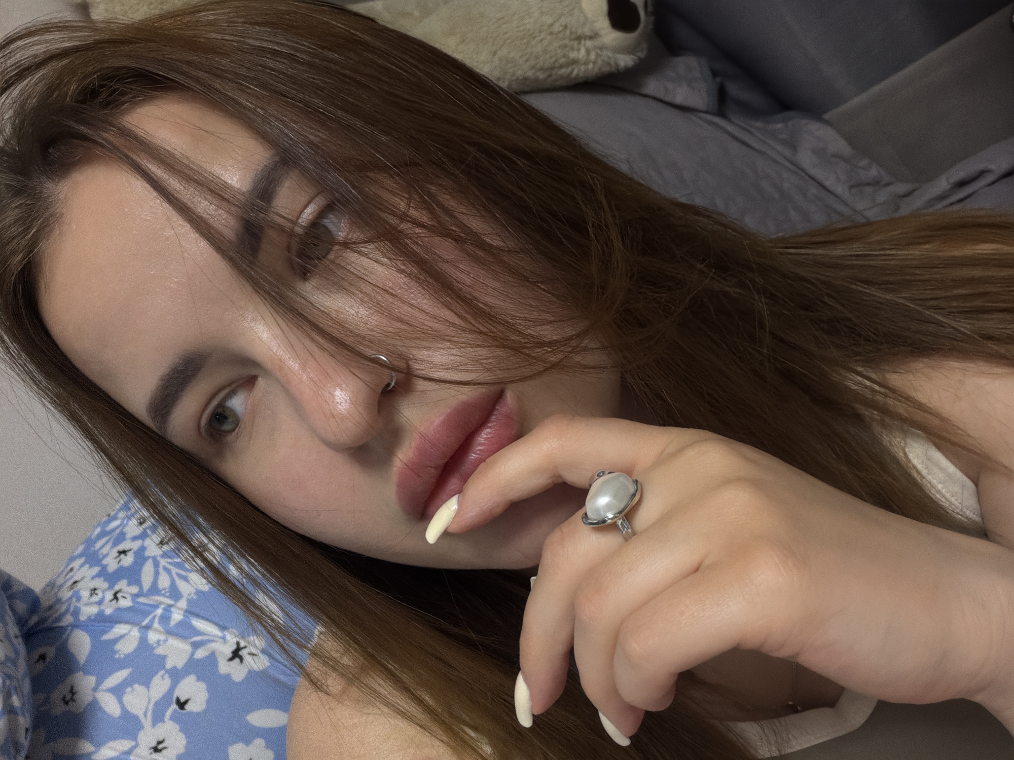 A young woman with light skin and long brown hair lies on a bed. Her head is tilted towards the camera, with some hair falling across her face. She has a nose ring and a large ring with a pearl-like gem on her right hand, which is near her mouth. She appears to be resting on pillows. The bed has a blue floral patterned pillowcase. In the background, a gray blanket and a stuffed animal are visible. The image was not taken in the city of Krasnodar, Russia, because it is impossible to derive the location from the image.