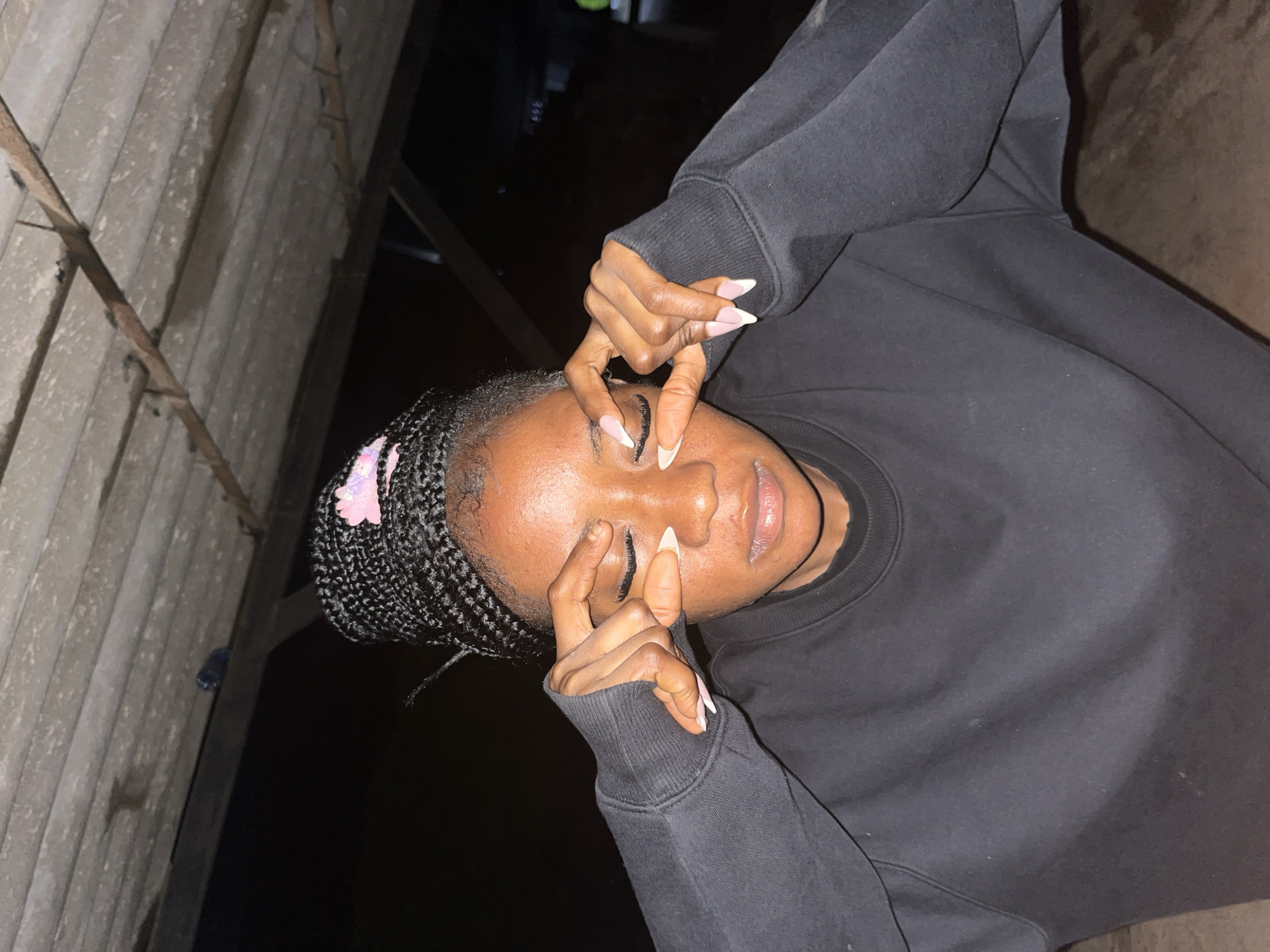 A young woman with braided hair, wearing a black sweatshirt, is pictured from the chest up. She is outdoors under a wooden ceiling, possibly in a covered area. Her eyes are closed, and her fingers are placed over them, obscuring her vision. The scene appears to be dimly lit, suggesting it might be evening or nighttime. The background is dark, with some indistinct structures visible. The woman's nails are painted with a French manicure. The location is identified as Jos, Nigeria.