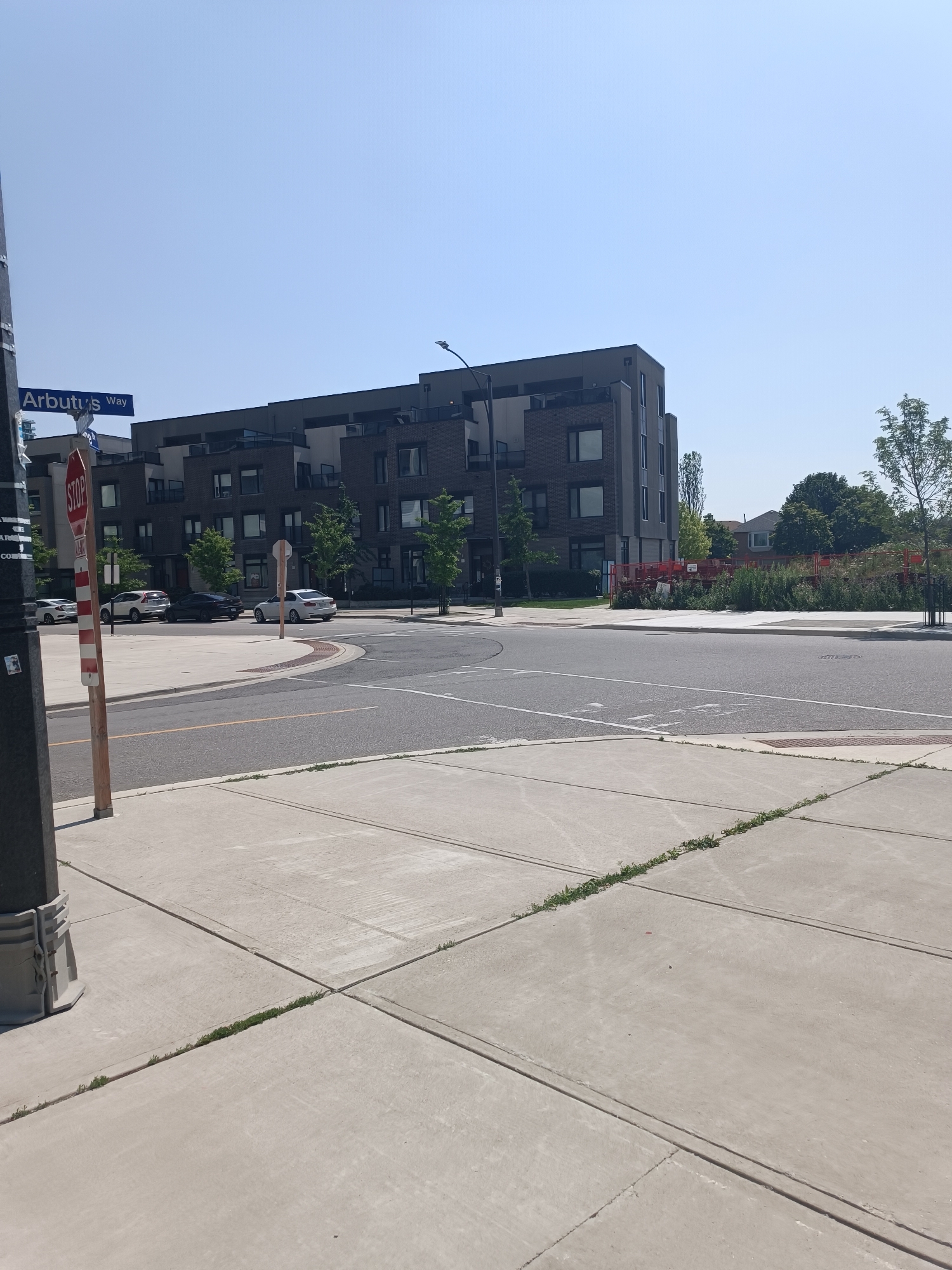 The image depicts an outdoor scene at a street intersection in Mississauga, Canada, on a clear day under a pale blue sky. In the foreground, concrete sidewalks with expansion joints and some sparse grass growing in them are visible. A dark pole with a metal base stands on the left.
The intersection features a curved road with painted lane markings and a concrete median. A red and white striped pole with a STOP sign is prominently placed on the sidewalk. Several cars are parked on the side of the road.
In the middle ground, a modern multi-story building with dark brick and gray siding, featuring numerous windows and balconies, is situated. To the right of the building, there is a patch of greenery with trees and what appears to be a construction site with red fencing and equipment.
In the background, a residential house with a sloped roof is visible behind the trees. The overall environment suggests a suburban or urban residential area undergoing some development. The street sign indicates "Arbutus Way."