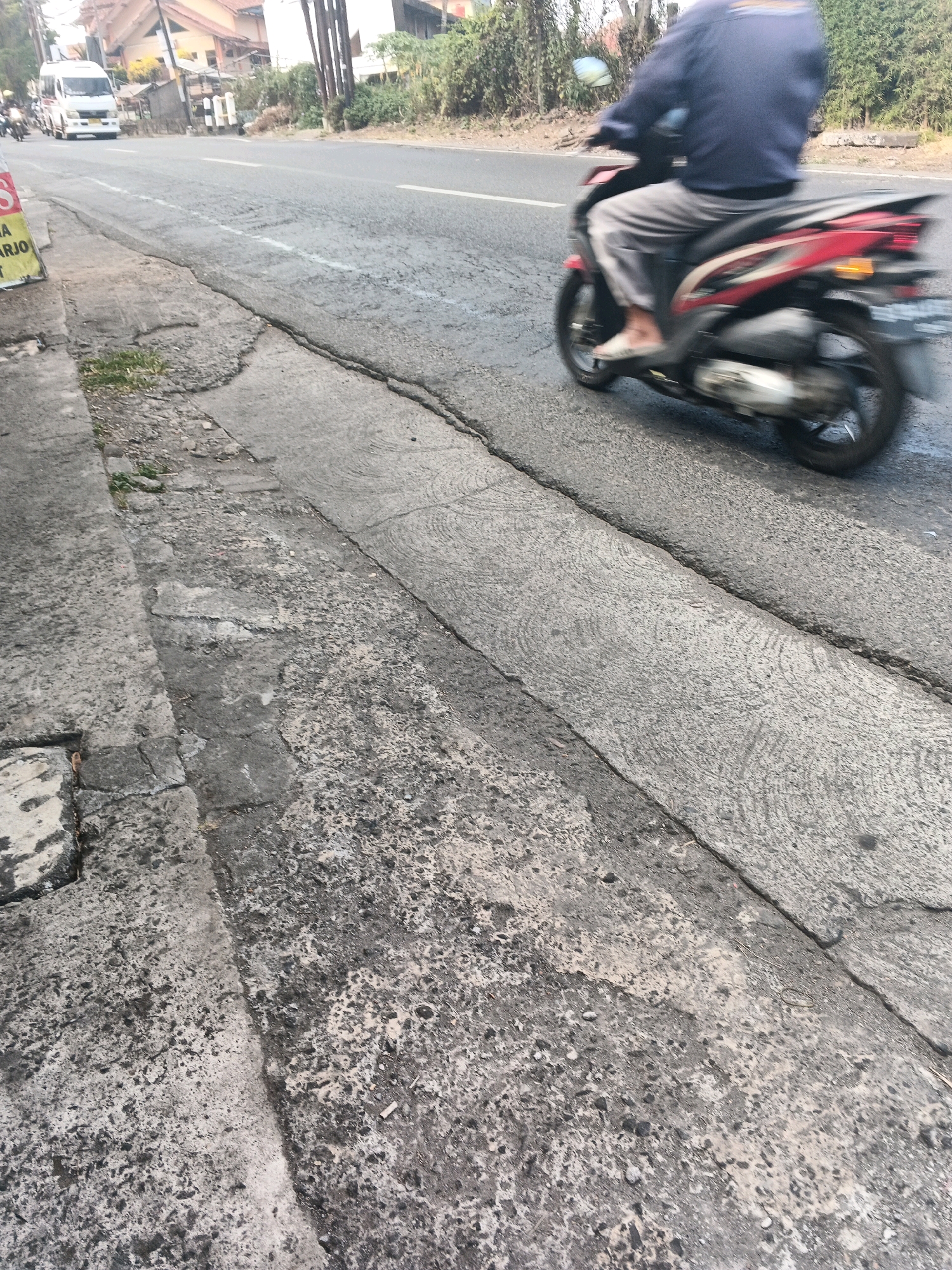 The image depicts a street scene in Ungaran, Indonesia. A person is riding a red and black motorcycle with motion blur, indicating movement. The rider is wearing a dark blue long-sleeved shirt and light gray pants. They are wearing light-colored footwear. The motorcycle is in motion on a paved road, which has several cracks and an uneven surface. To the left of the road is a concrete sidewalk with some vegetation growing through it. In the background, a white van is visible, and the scene suggests an urban or semi-urban environment with buildings and trees.