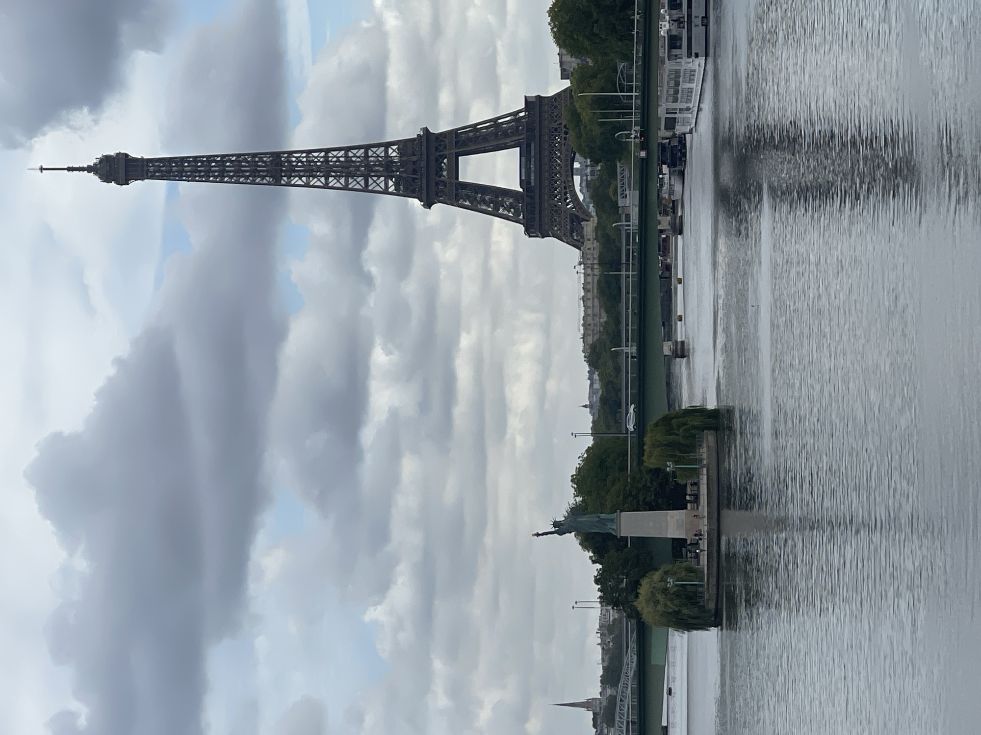 The image captures a view of the Seine River in Paris, France, on a cloudy day. Prominently featured is the Eiffel Tower in the background, a tall iron lattice tower. In the foreground, on a small island in the river, stands a replica of the Statue of Liberty, holding her torch aloft. A green bridge spans the river in the mid-ground, with vehicles visible on it. Several boats, including what appears to be a river cruise boat, are docked or moving along the river. The water reflects the overcast sky and the surrounding structures. Trees and buildings line the riverbanks, with a church spire visible in the distance to the left. The scene suggests a calm day with moderate cloud cover.