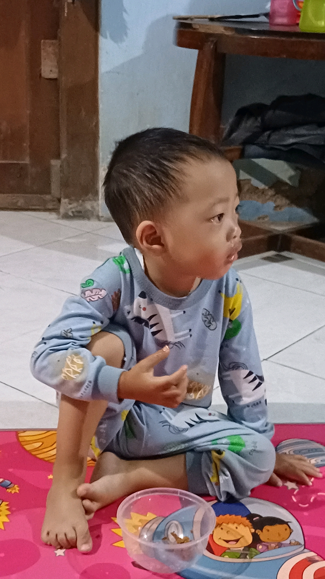 A young East Asian child, appearing to be around 2-3 years old, is seated cross-legged on a pink mat with a space-themed print. The child is wearing light blue pajamas with a pattern of zebras and other cartoon animals. Their right arm is bent, with their hand resting on their lap, and their left arm is also bent, with fingers extended. A small, clear plastic bowl containing some brown material is positioned to the child's right. The background shows a tiled floor and a wooden structure, suggesting an indoor domestic setting, potentially in Wonokerso, Indonesia. The lighting is subdued, with shadows cast on the wall behind the child. The child's gaze is directed to the right, with their lips slightly parted.