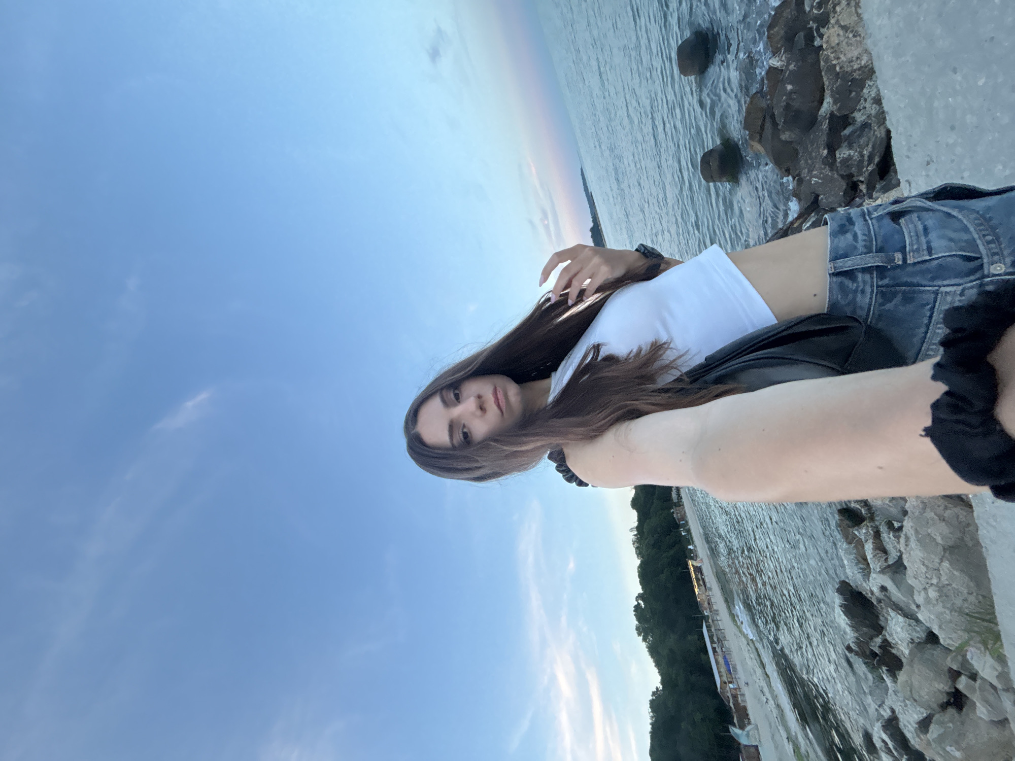 A young woman with long brown hair and a white cropped t-shirt and blue jeans is taking a selfie on a rocky shore by the sea, with a beach and trees in the background. The location is Varna, Bulgaria, likely near the Sea Garden, a large park with a beach area. The sky is blue with some wispy clouds. The sea is calm with small waves.