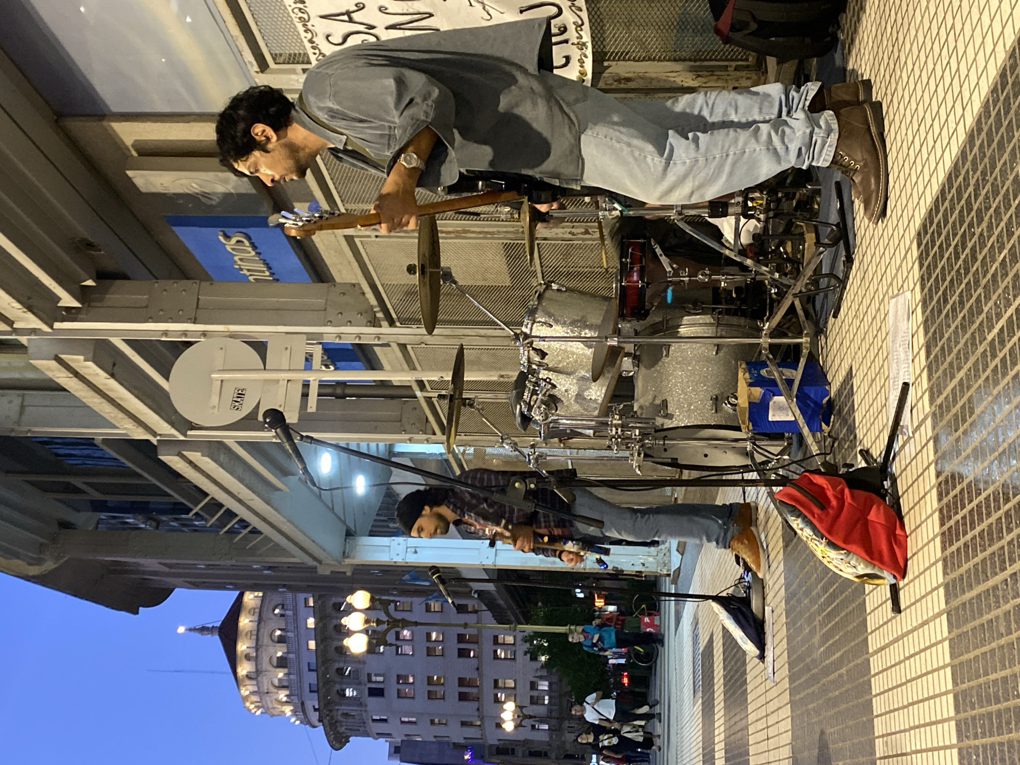 A long shot depicts two street musicians performing outdoors in Buenos Aires, Argentina. The camera angle is rotated approximately 90 degrees clockwise, providing a unique perspective.
The upper musician wears a gray shirt, light blue jeans, and brown boots. He plays the drums.
Below him, the second musician wears a plaid shirt, light blue jeans, and brown boots. He is positioned behind a microphone and plays a guitar.
The background includes a building with illuminated windows and streetlights, visible under an elevated structure bearing the sign "gentinas". A small crowd gathers near the bottom of the image.
Additional details include a drum set, musical equipment, and a red backpack placed on the ground. The musicians stand on a patterned tile sidewalk.