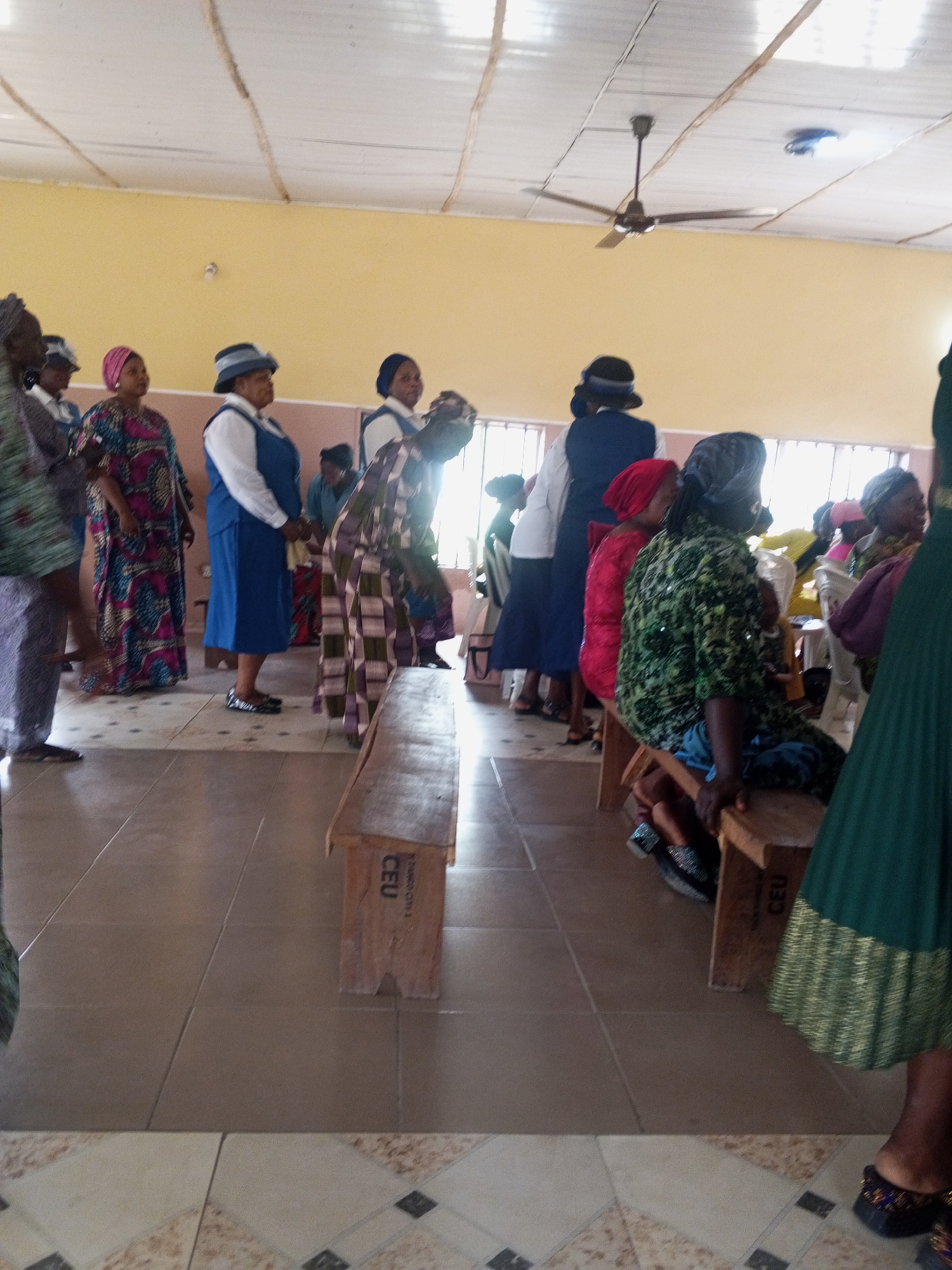 The image depicts an indoor gathering of predominantly women in Zango-ndaji, Nigeria. The setting appears to be a community hall or church, with a tiled floor and a suspended ceiling with exposed beams and a ceiling fan. The walls are painted a pale yellow.
In the foreground, several wooden benches are visible, one of which has the inscription "CEU" on its side. A woman in a dark green pleated skirt with gold trim is standing to the right, partially obscuring the view of the bench. Her feet, adorned with embellished dark shoes, are visible.
Further into the scene, a group of women are standing and seated. Several women are wearing patterned dresses and head coverings. One woman in the center is wearing a blue dress over a white shirt and a blue hat. Another woman to her left is in a vibrant, colorful patterned dress. Some individuals appear to be engaged in conversation or observing an event. The lighting suggests it is daytime, with natural light entering from windows or openings on the right side of the frame. The overall atmosphere appears to be one of a community event or religious service.