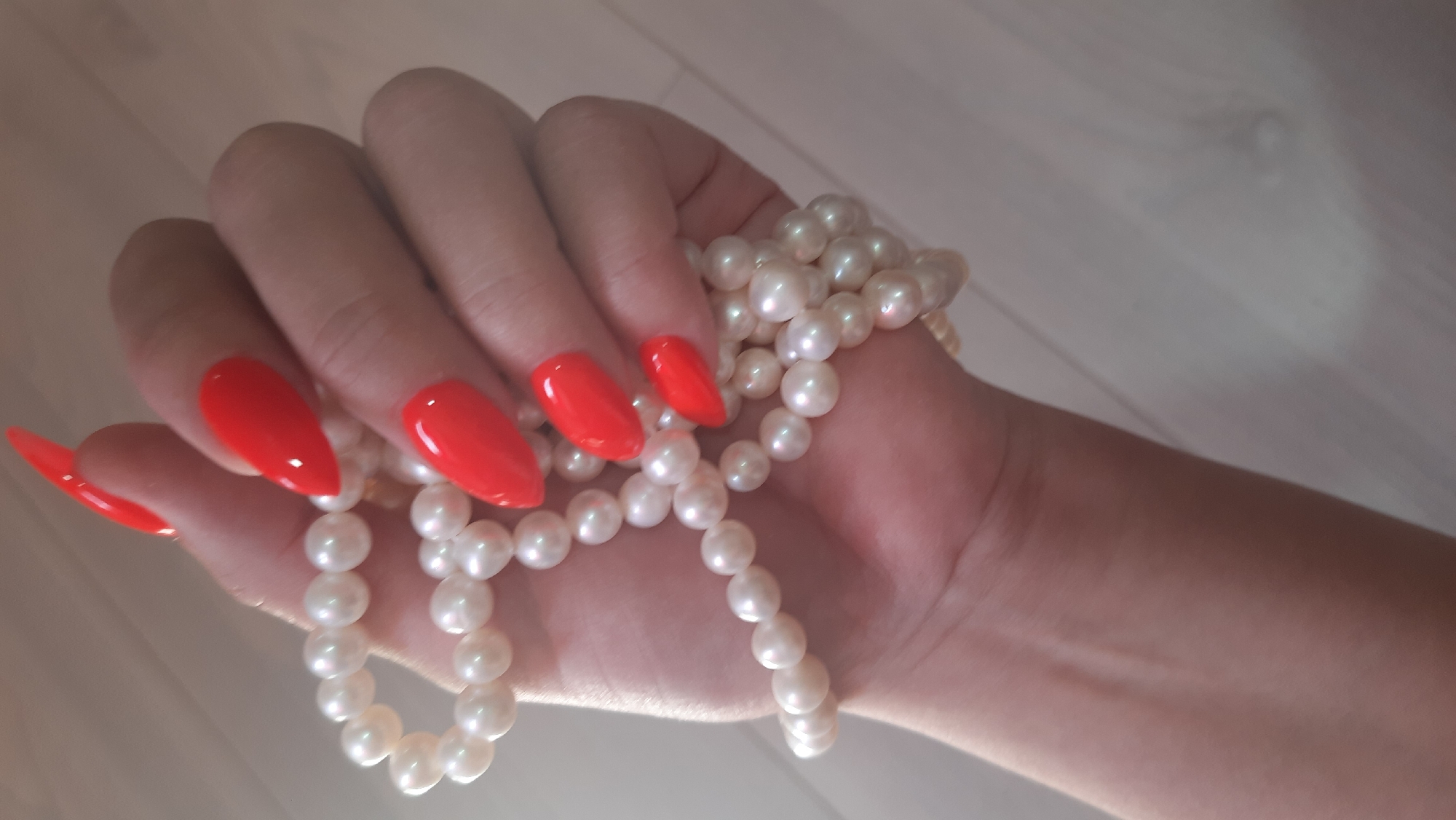 A close-up shot captures a hand adorned with bright red, almond-shaped nails holding a string of pearls. The hand is positioned over a light-colored, wood-grained surface, possibly a floor or table, with a blurred background. The pearls are small and white, with a subtle sheen. The lighting appears to be soft, casting gentle shadows and highlighting the textures of both the pearls and the nails. The image does not provide any information regarding a specific location in Desnogorsk, Russia.