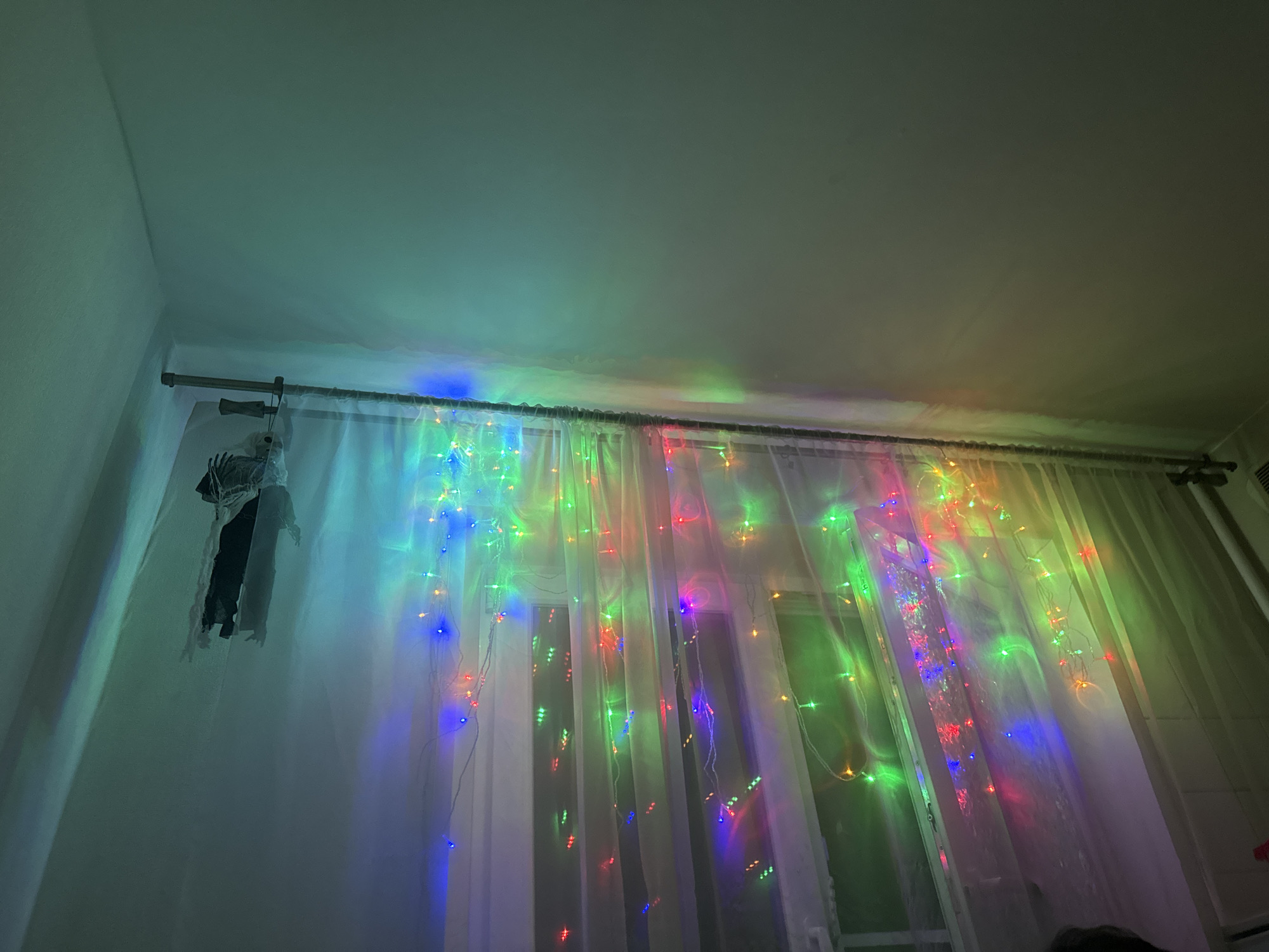 The image shows a room interior with a window covered by white sheer curtains. A string of colorful LED lights is draped across the curtains, casting a vibrant glow. The lights are red, green, blue, and yellow, creating a festive and playful atmosphere. On the left side of the image, hanging from the curtain rod, is a decorative ghost made of white fabric with a black body. The room appears to be lit by the colorful lights and possibly ambient room lighting, as the overall lighting is dim. The ceiling is illuminated by the lights, showing a gradient of colors. The scene suggests a celebration or holiday decoration, possibly for Halloween given the ghost. The location is Moscow, Russia. No text is visible in the image.