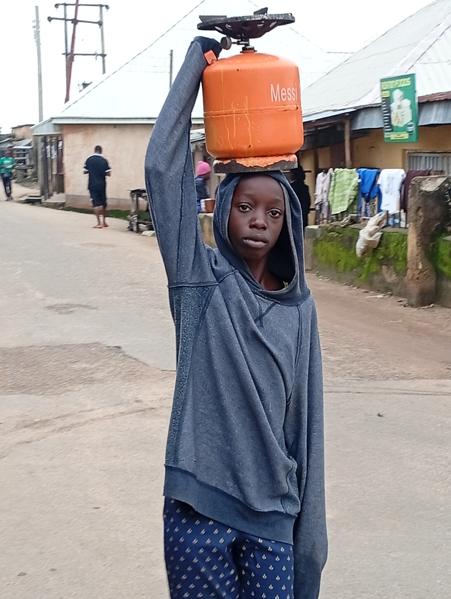 A young person with dark skin and black hair is shown from the chest up, balancing an orange propane tank on their head. The tank has the word "Messi" written on it in white letters. They are wearing a grey hooded sweatshirt and blue patterned pants. The person is standing on a paved street with a slight incline. In the background, there are several single-story buildings with corrugated metal roofs. Power lines and a utility pole are visible in the upper left corner. A green sign with text that is difficult to read is hanging on one of the buildings. Clothes are hanging on a line in front of another building. The scene appears to be in a residential area of Jos, Nigeria.